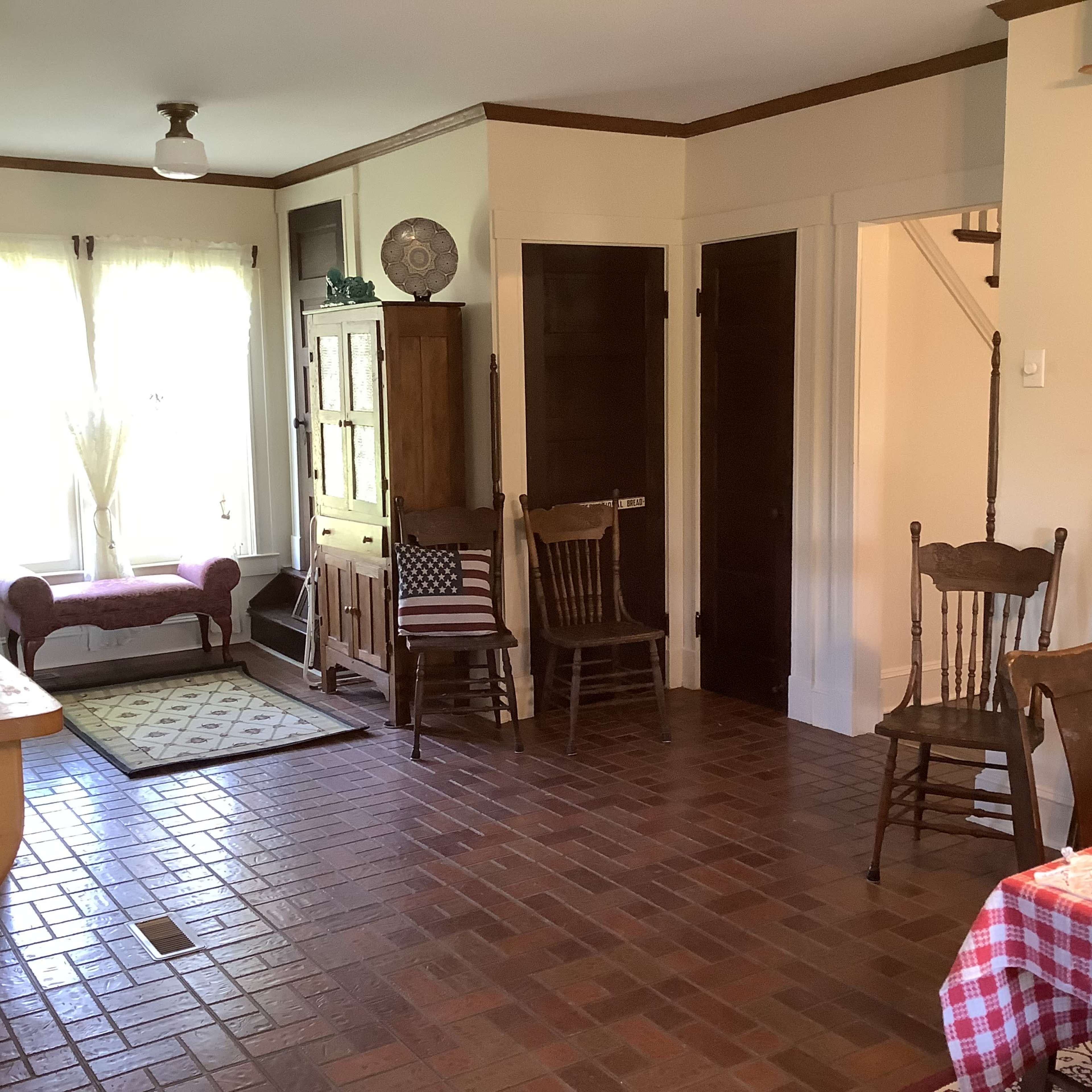 The image shows a cozy living space with brick flooring, a pink chaise lounge near a window, and several wooden chairs arranged around the area.