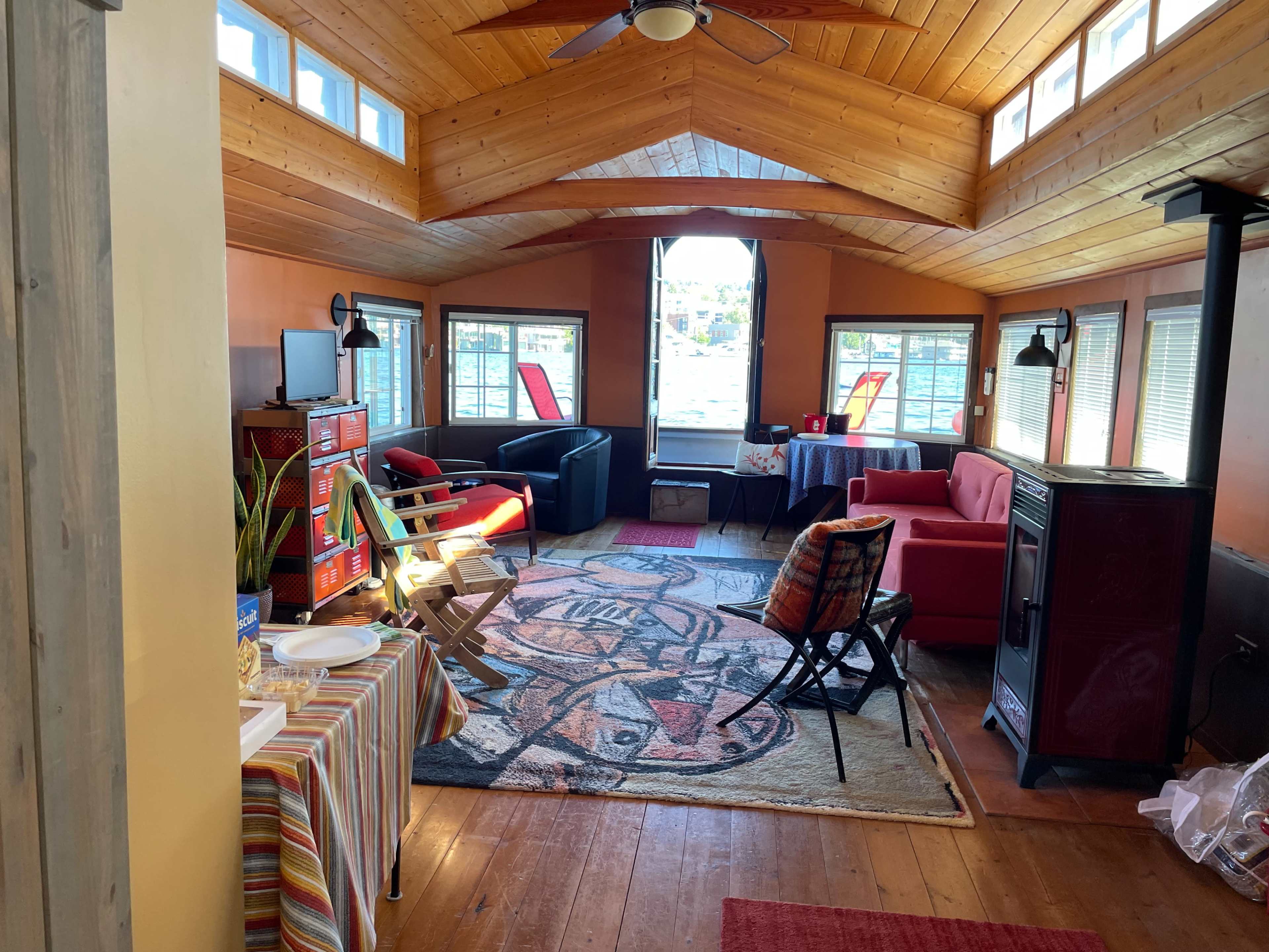 The image shows a cozy sunlit room with wooden walls and ceiling, featuring seating areas with chairs and sofas, a table, and a stove.