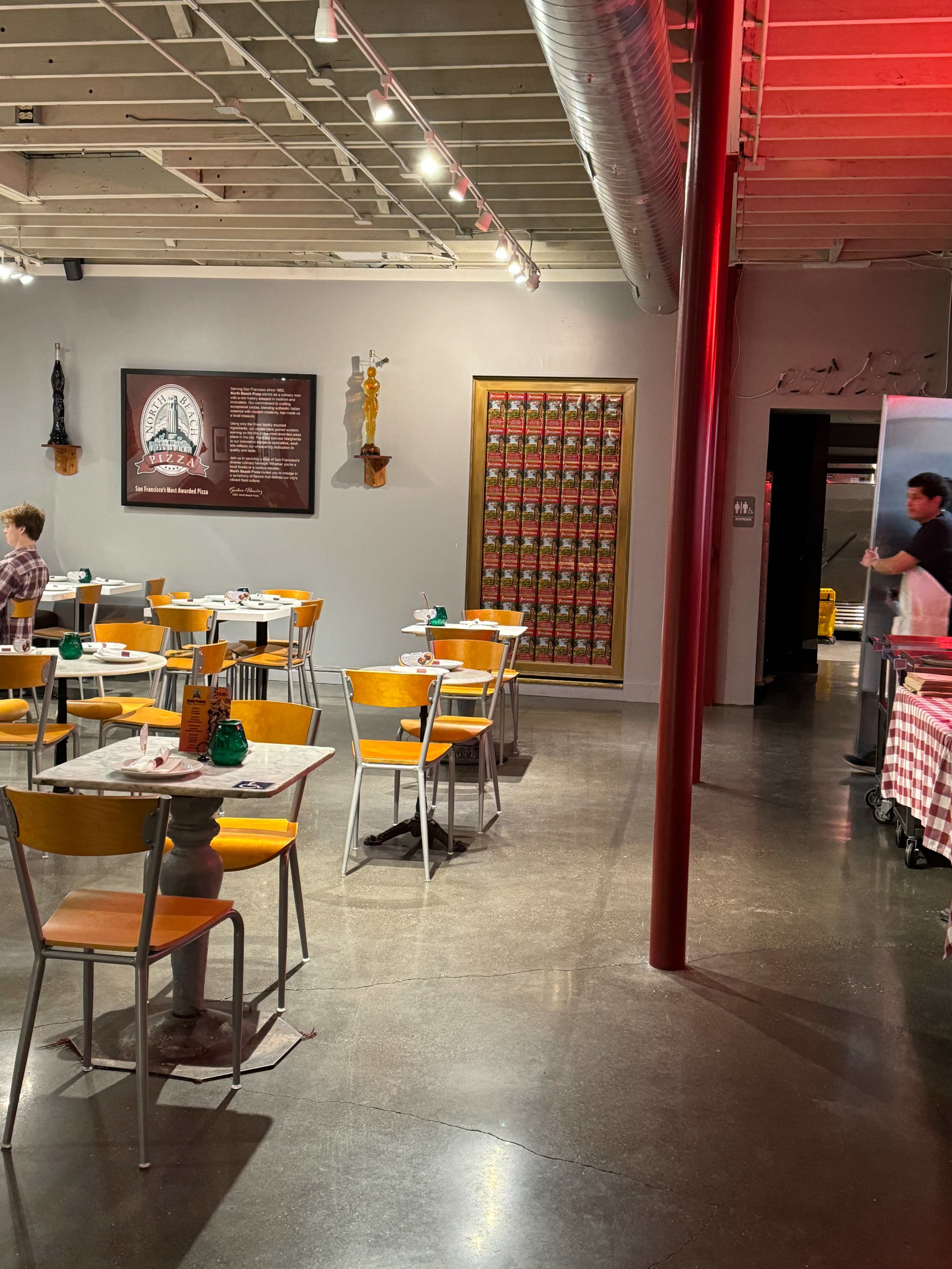 A dining area in a restaurant featuring wooden tables and chairs, with a wall of decorative cans and a kitchen entrance visible at the far end.