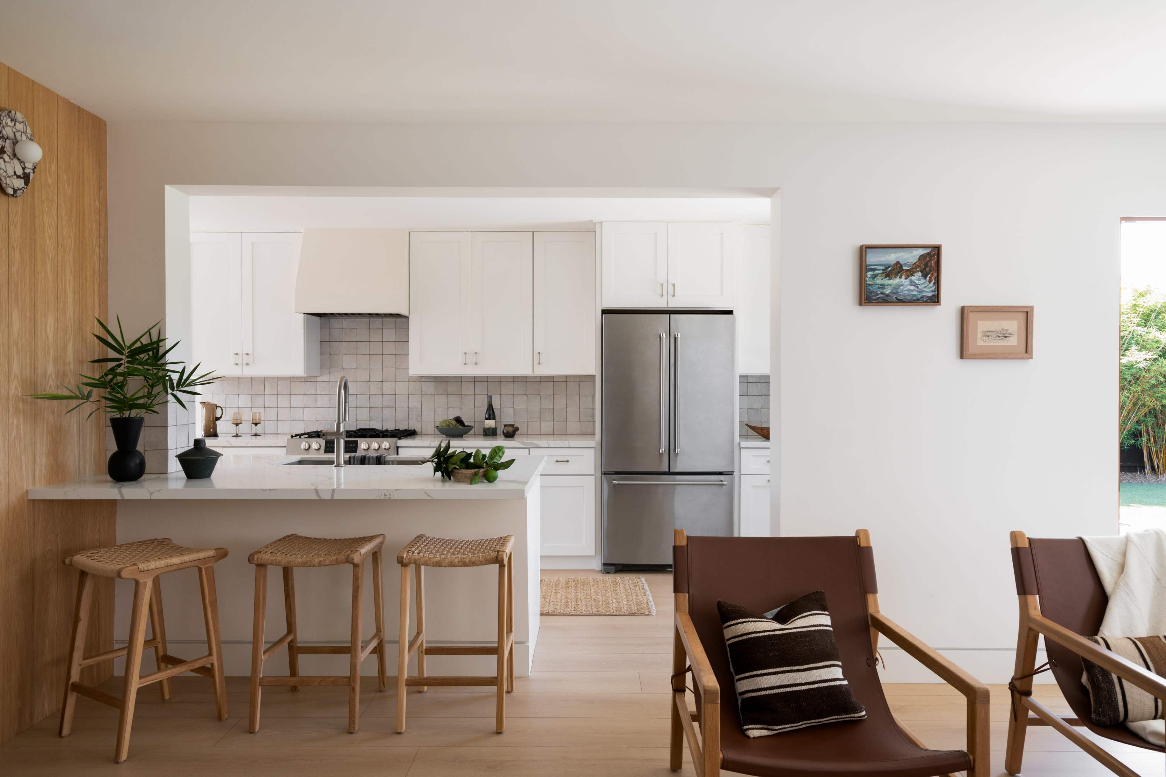 The image shows a modern kitchen with white cabinetry, stainless steel appliances, and a bar area with wooden stools, adjacent to a living space featuring two wooden chairs.