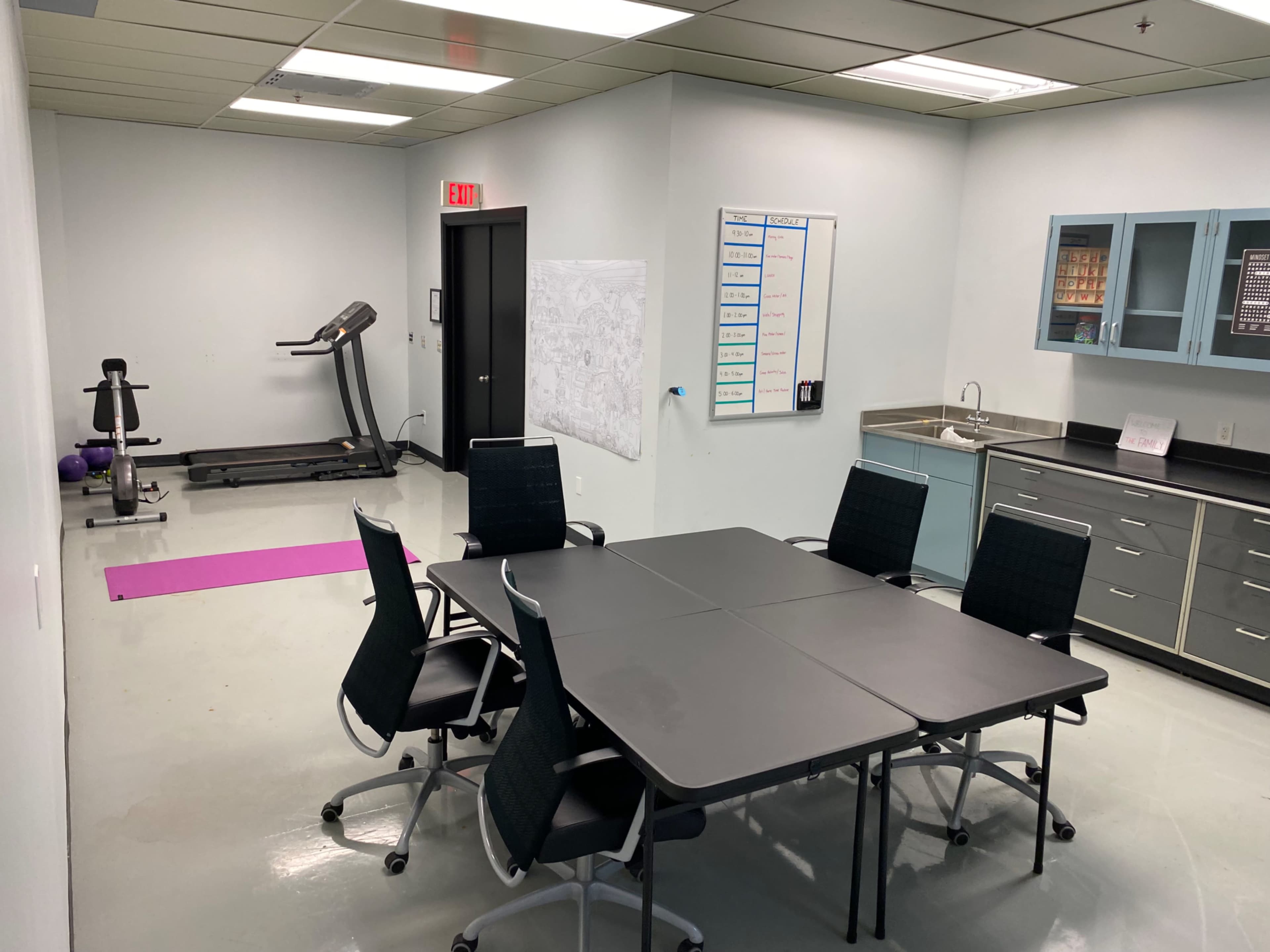 A well-lit room featuring a conference table surrounded by six chairs, a treadmill in the corner, and a kitchenette area with cabinets and a sink.