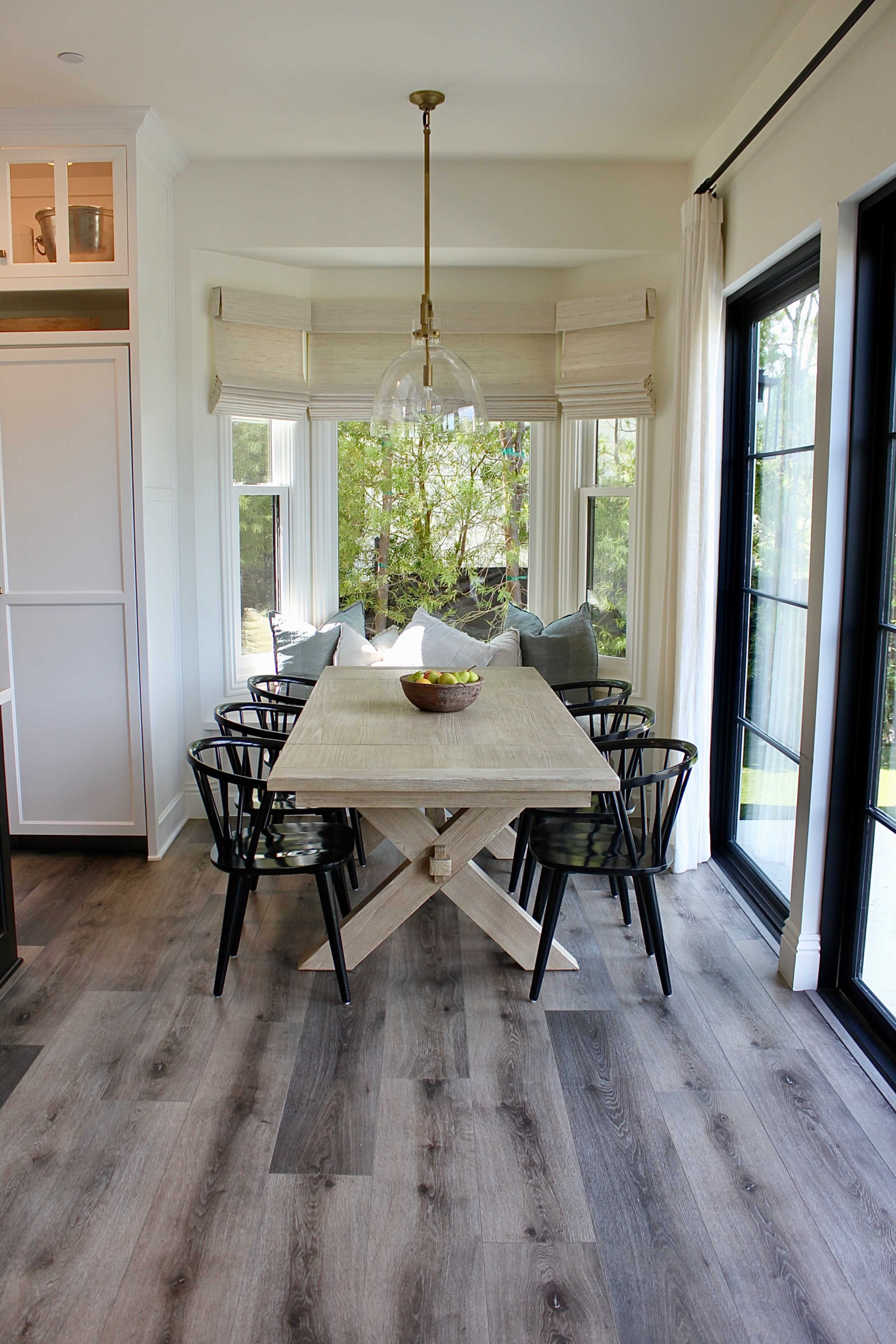 A modern dining area features a light wooden table surrounded by black chairs, with large windows offering a view of greenery outside.