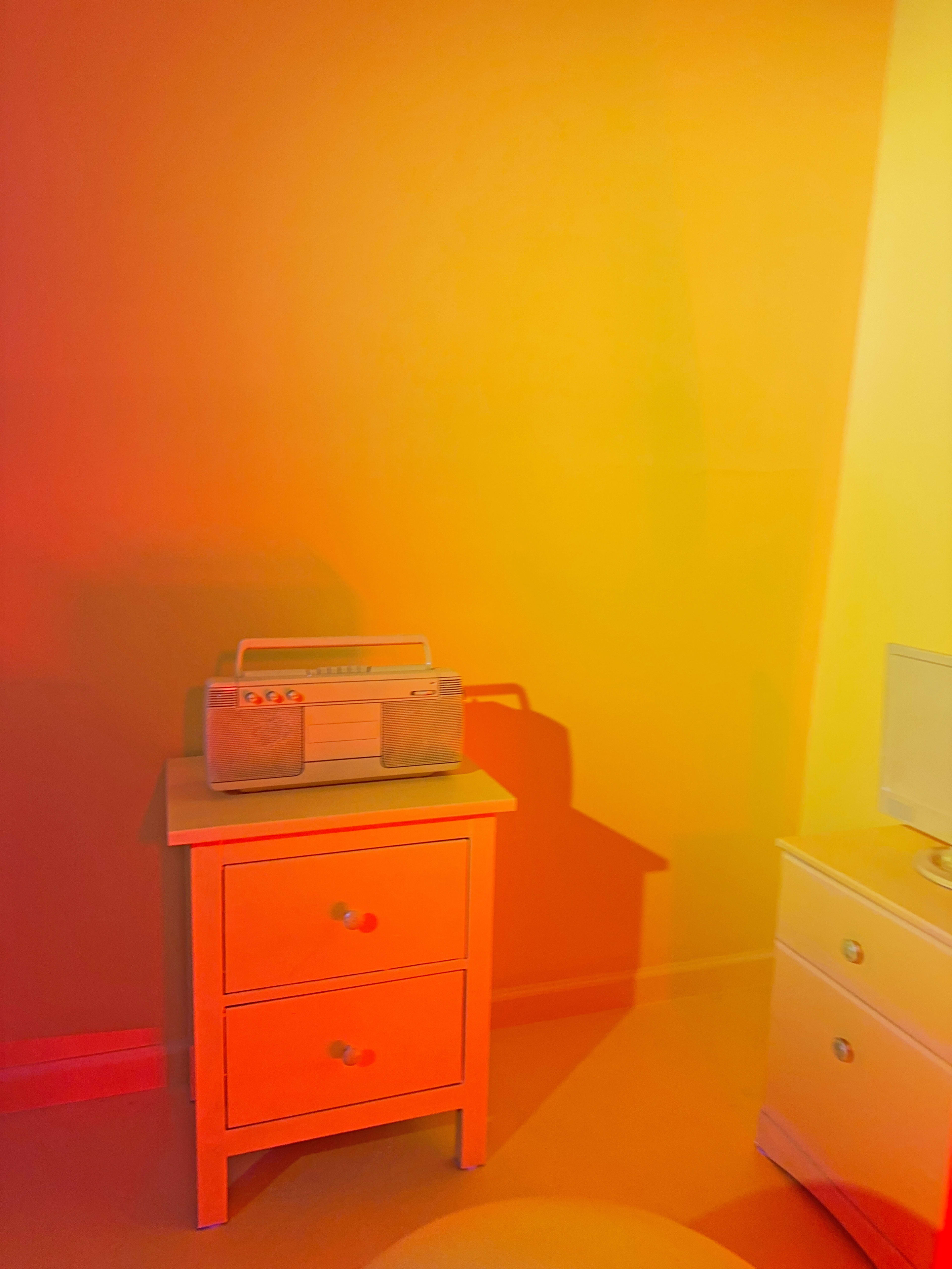 A small room features a wooden bedside table with two drawers, holding a vintage radio, set against brightly colored yellow and orange walls.