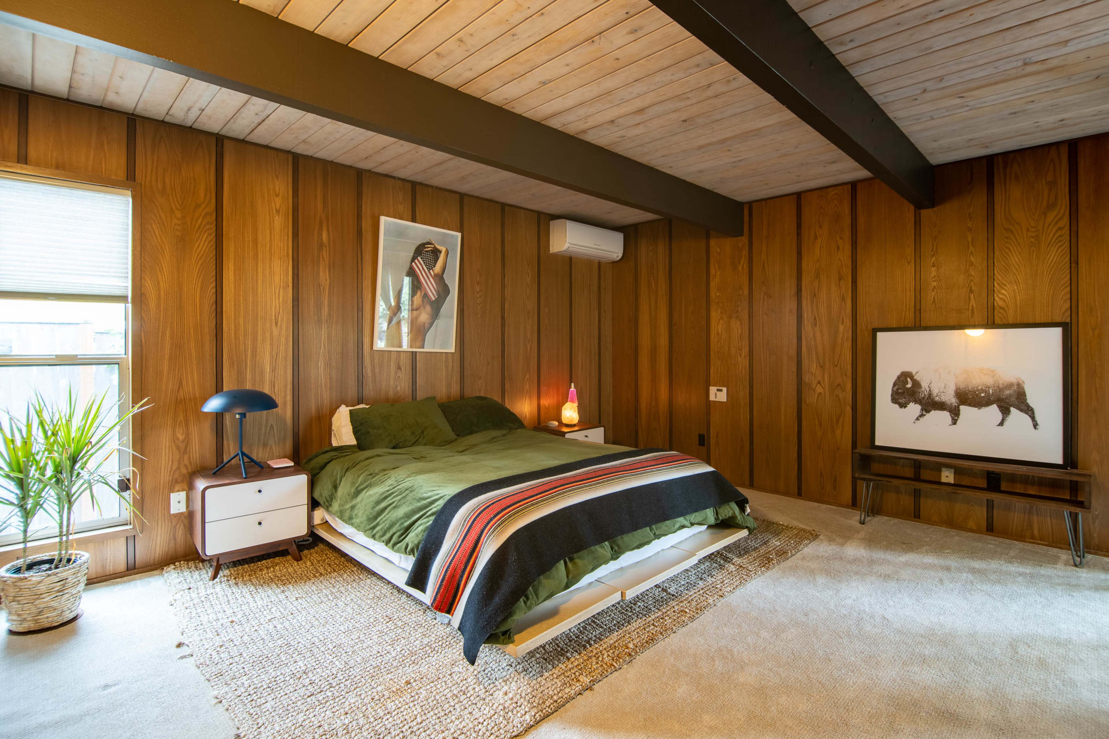 A wooden-paneled bedroom with a green bedspread, a lamp, a plant by the window, and artwork featuring a buffalo on the wall.