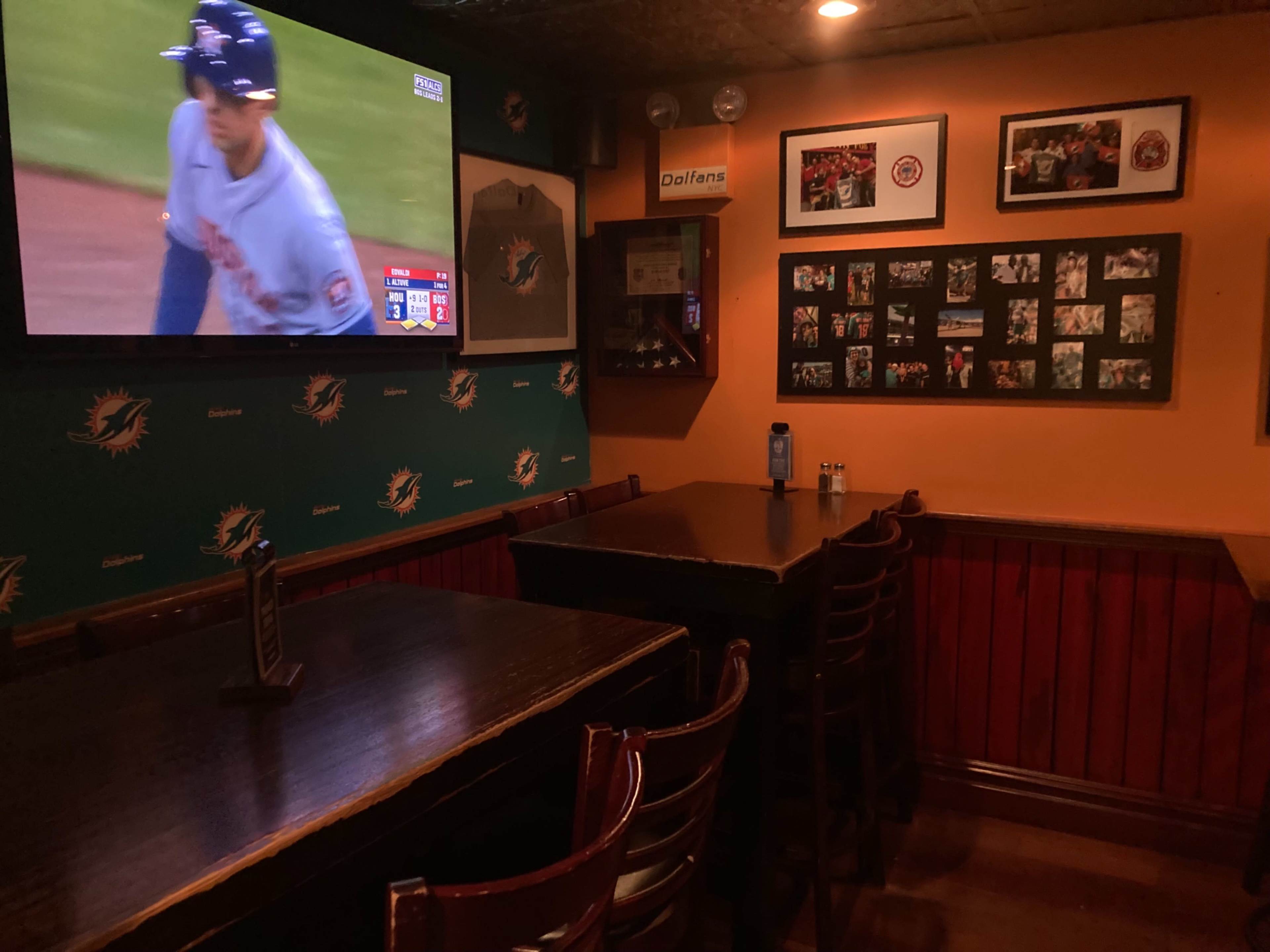 A dimly lit bar features a large television displaying a baseball game, with empty wooden tables and Dolphins-themed decor on the walls.