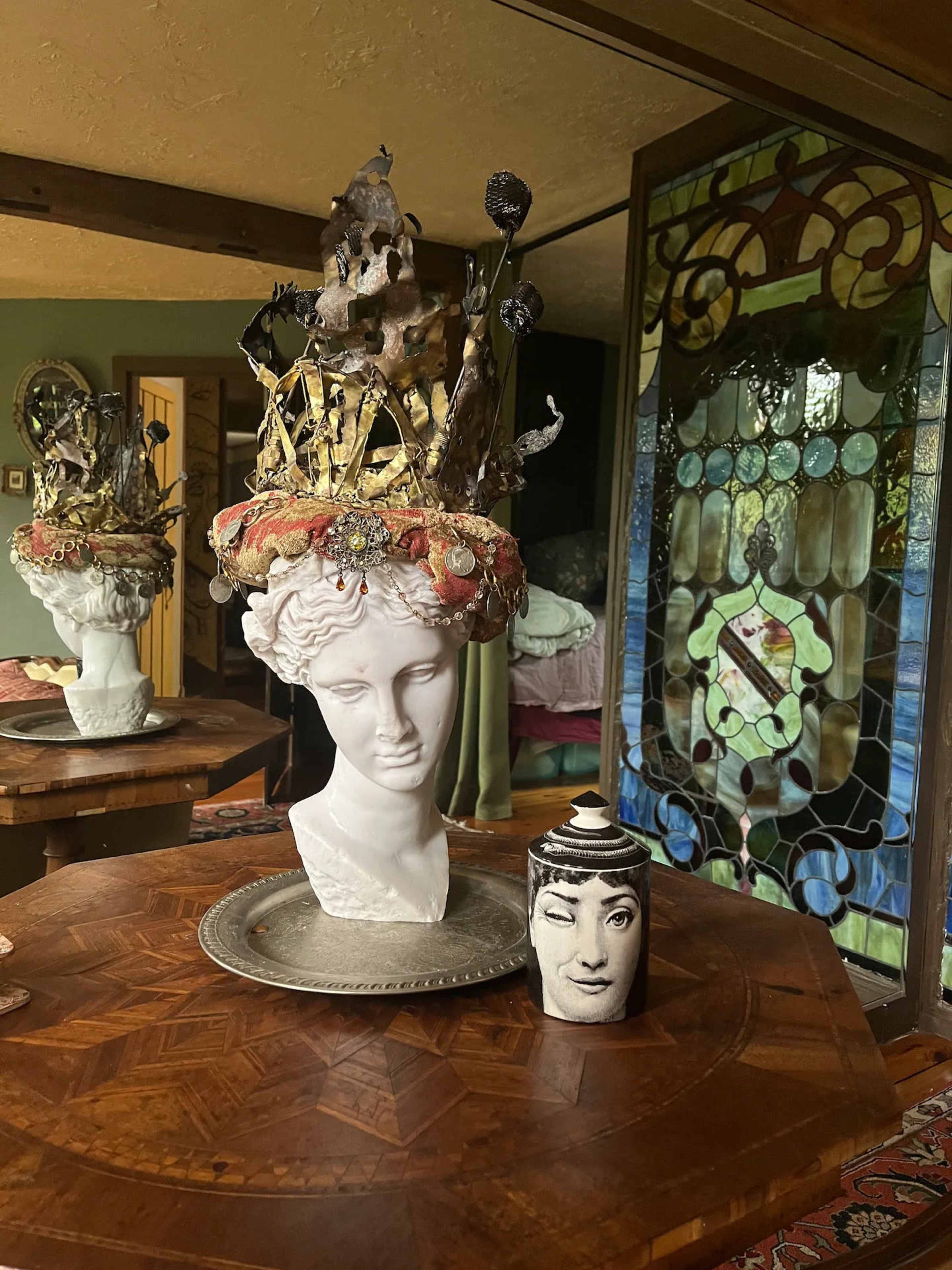A decorative bust with an elaborate crown sits on a wooden table beside a vintage vase featuring a graphic face, with stained glass in the background.