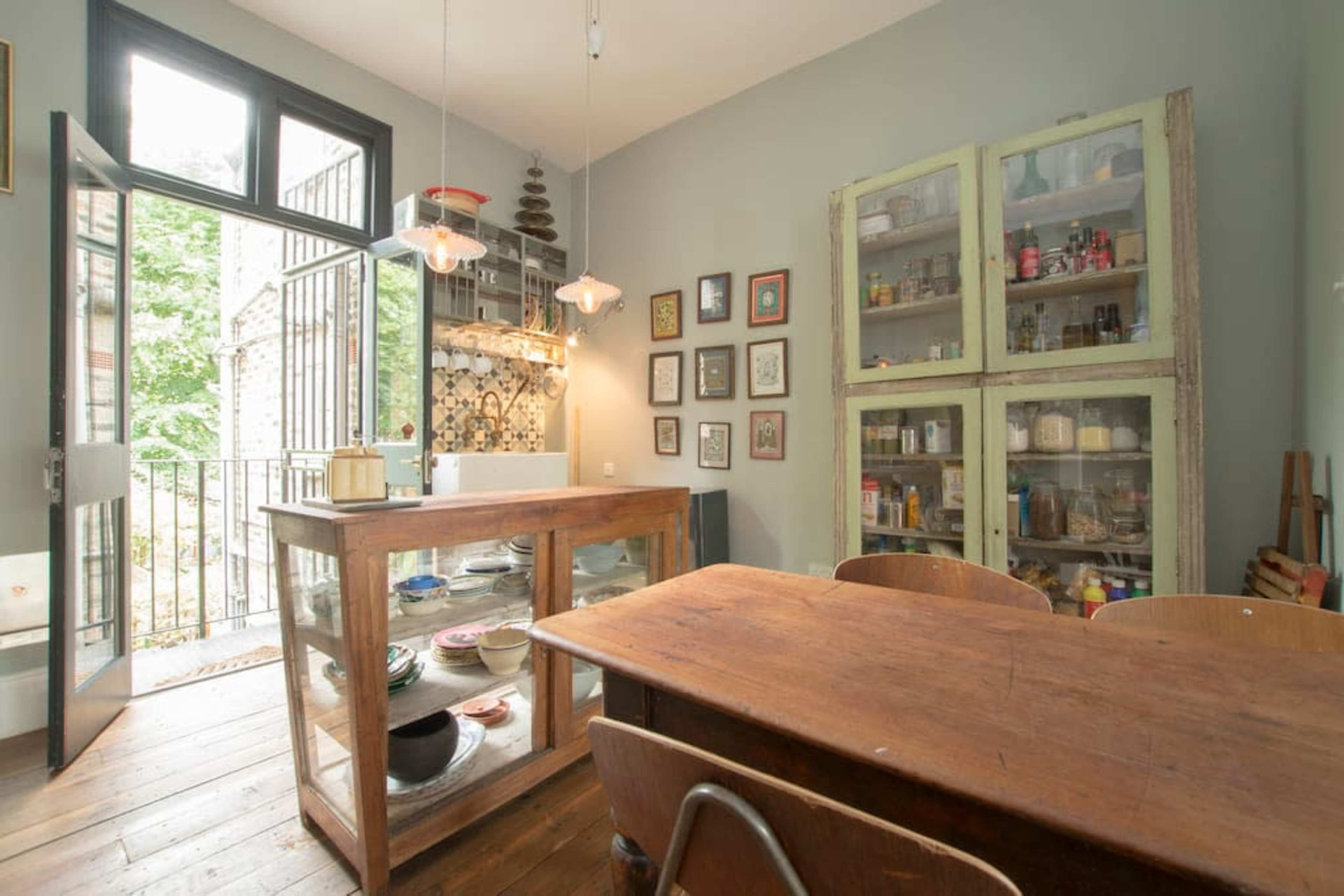 A kitchen with a wooden table, an open doorway leading to a balcony, and a cabinet filled with jars and dishes.