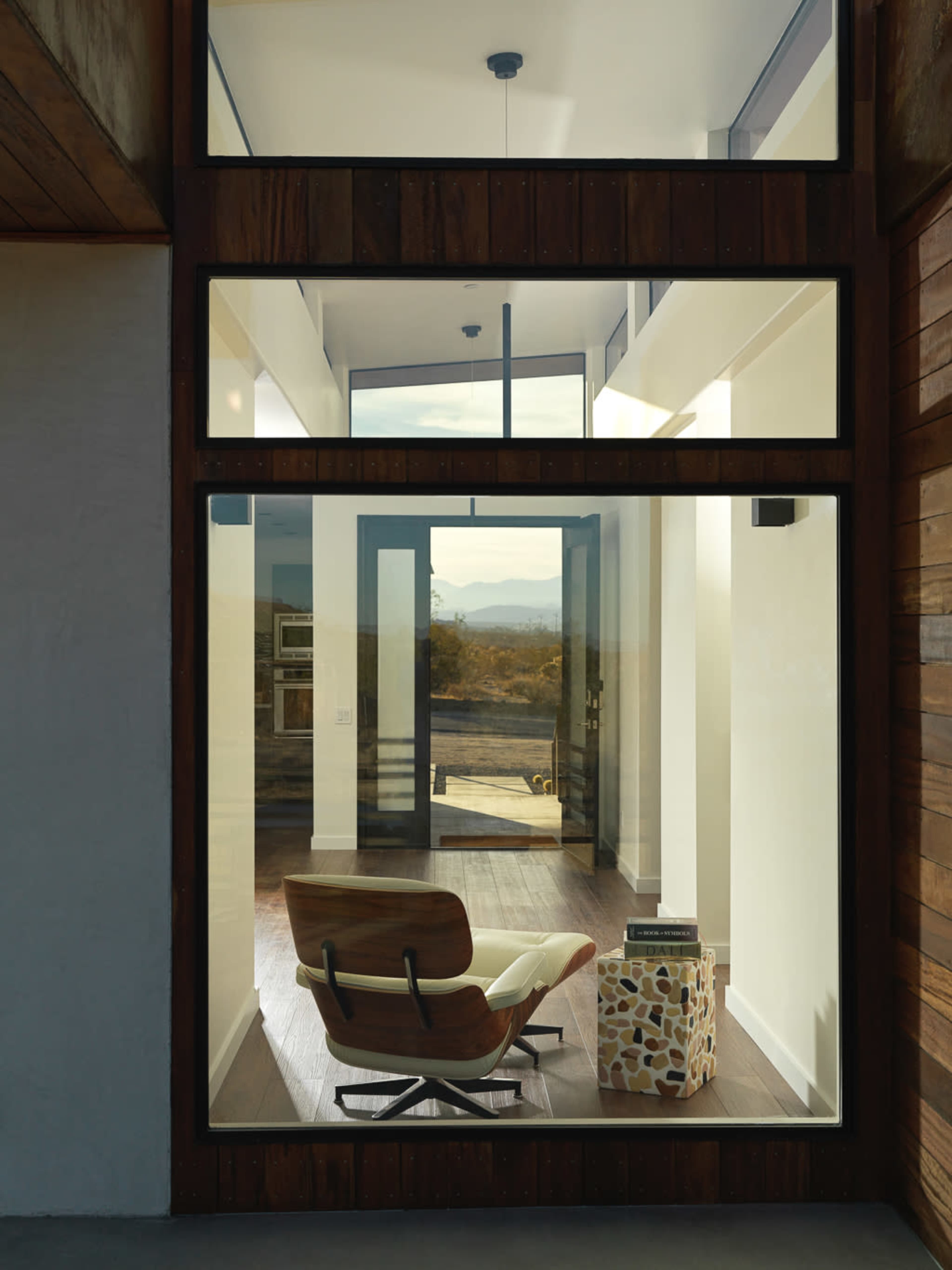 A modern interior space features a wooden chair and a patterned side table, with large glass windows showcasing a mountain view in the background.