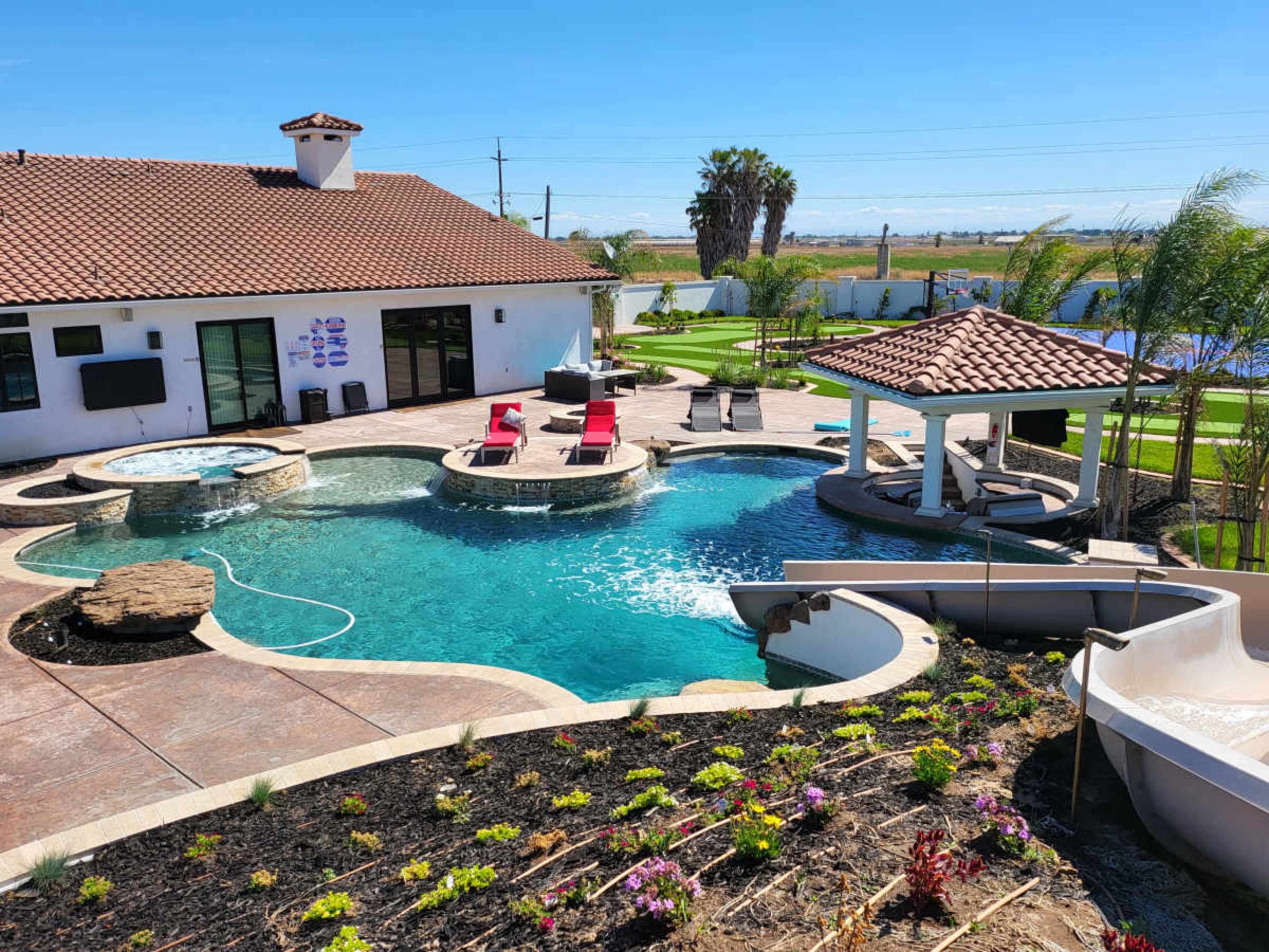 The image shows a modern backyard with a pool featuring a waterfall and a slide, surrounded by landscaped gardens and a gazebo.