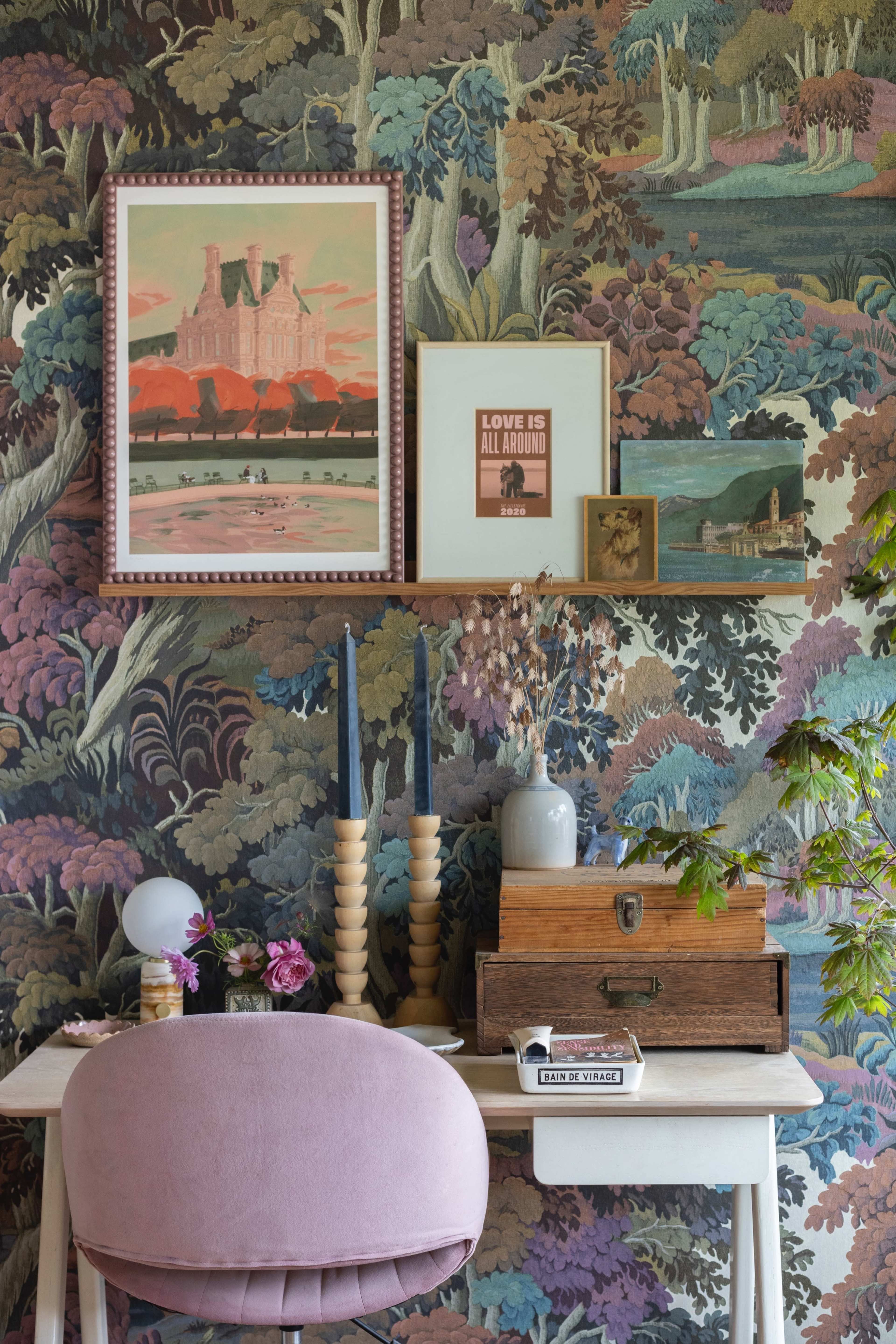 A decorative wall features patterned wallpaper with a shelf displaying framed art and small decorative items above a light-colored desk with a pink chair, candles, and a wooden box.