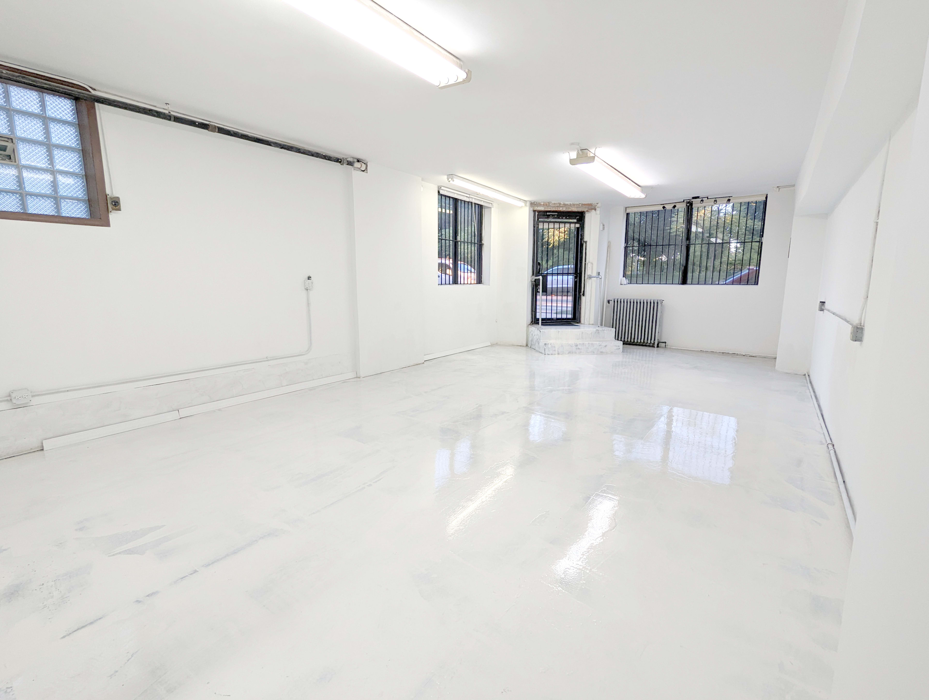 A spacious, empty room with white walls and a glossy white floor, featuring multiple windows with metal bars.