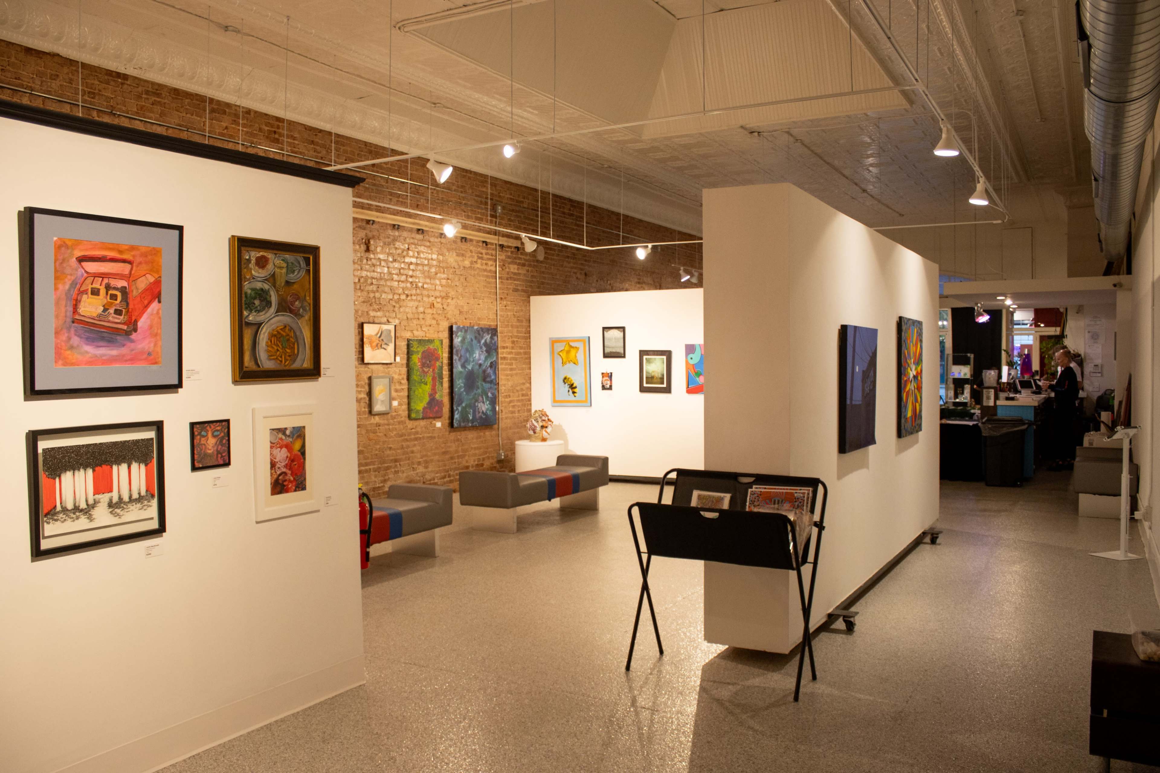 The image shows an art gallery featuring various framed artworks displayed on white walls and a brick backdrop, with seating and a reception area visible in the background.