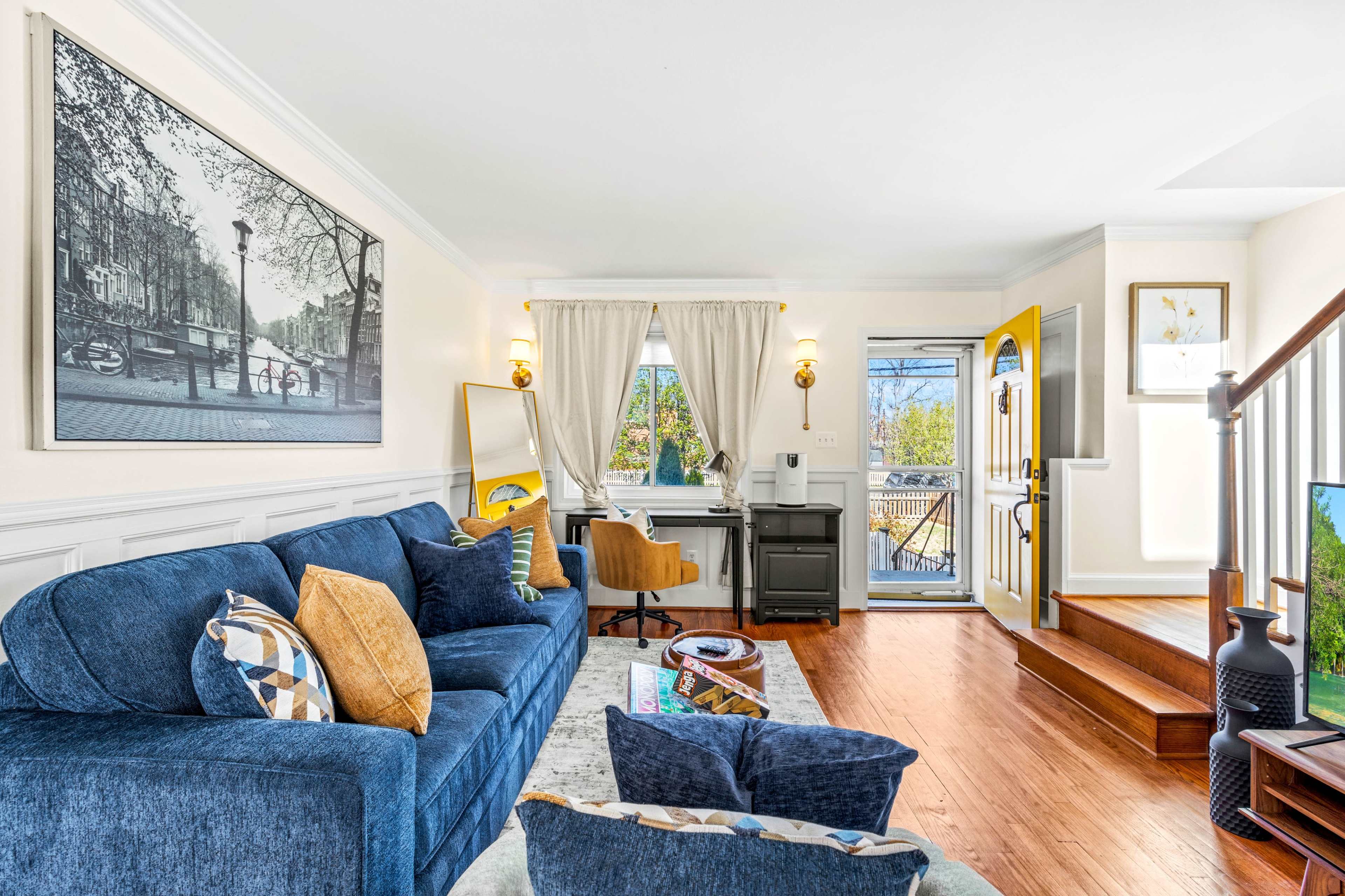 A cozy living room features a blue sofa, a small workspace by a window, and a staircase leading to an upper level.