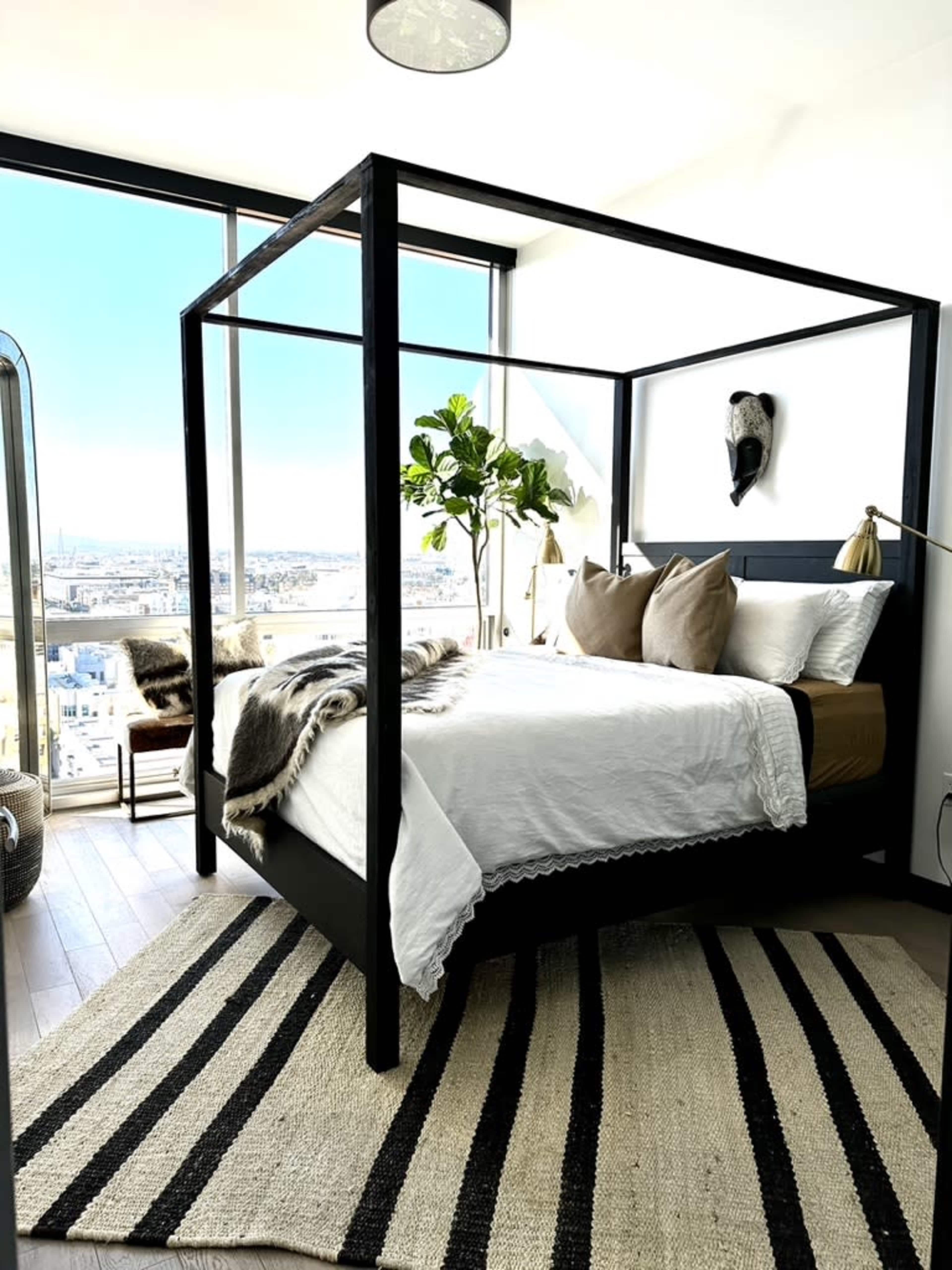 A modern bedroom features a black four-poster bed with a white bedding set, a large window offering a city view, and a striped rug on the wooden floor.
