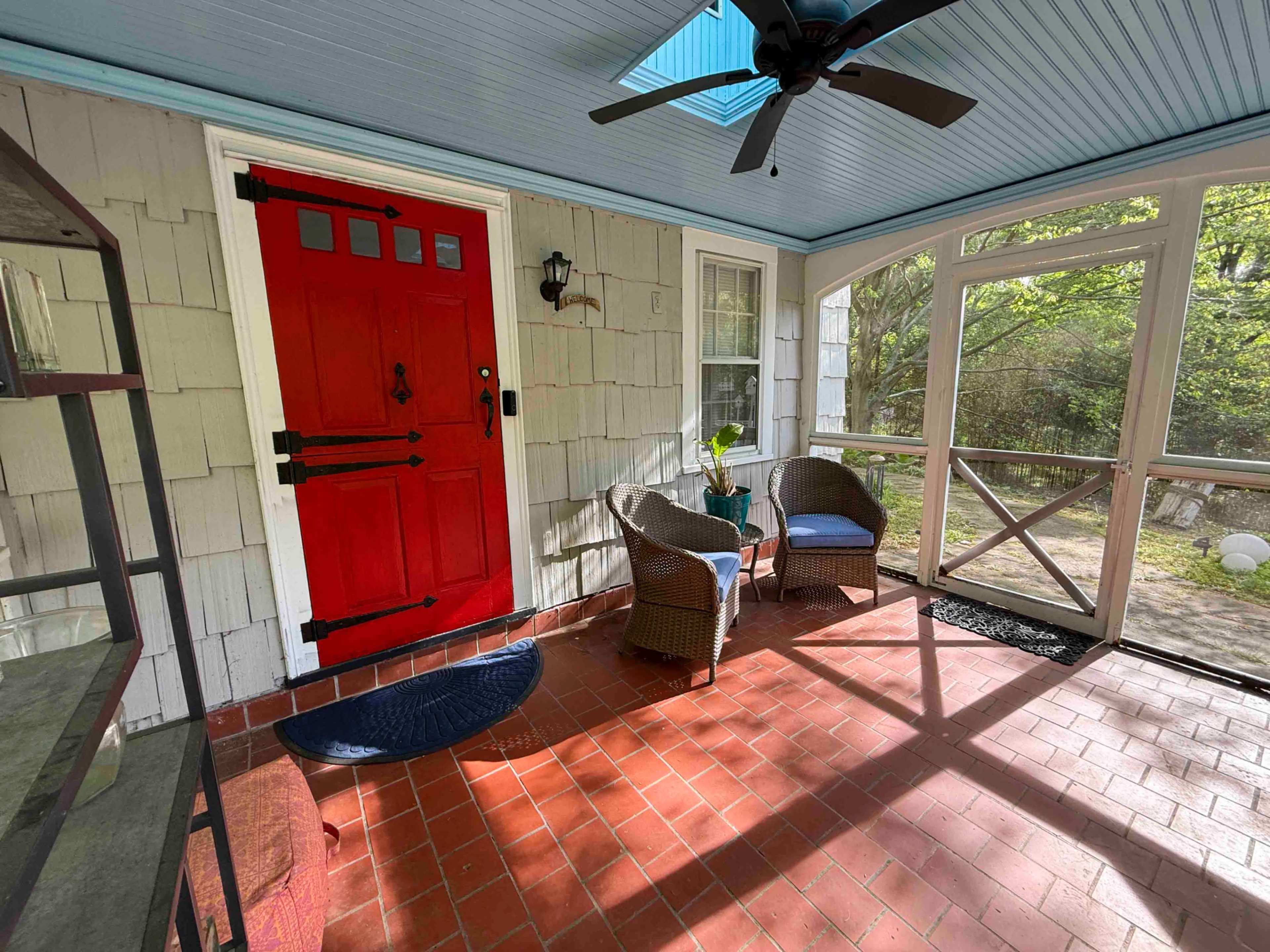 The image shows a screened porch with a red front door, a pair of wicker chairs, and a small table on a tiled floor.