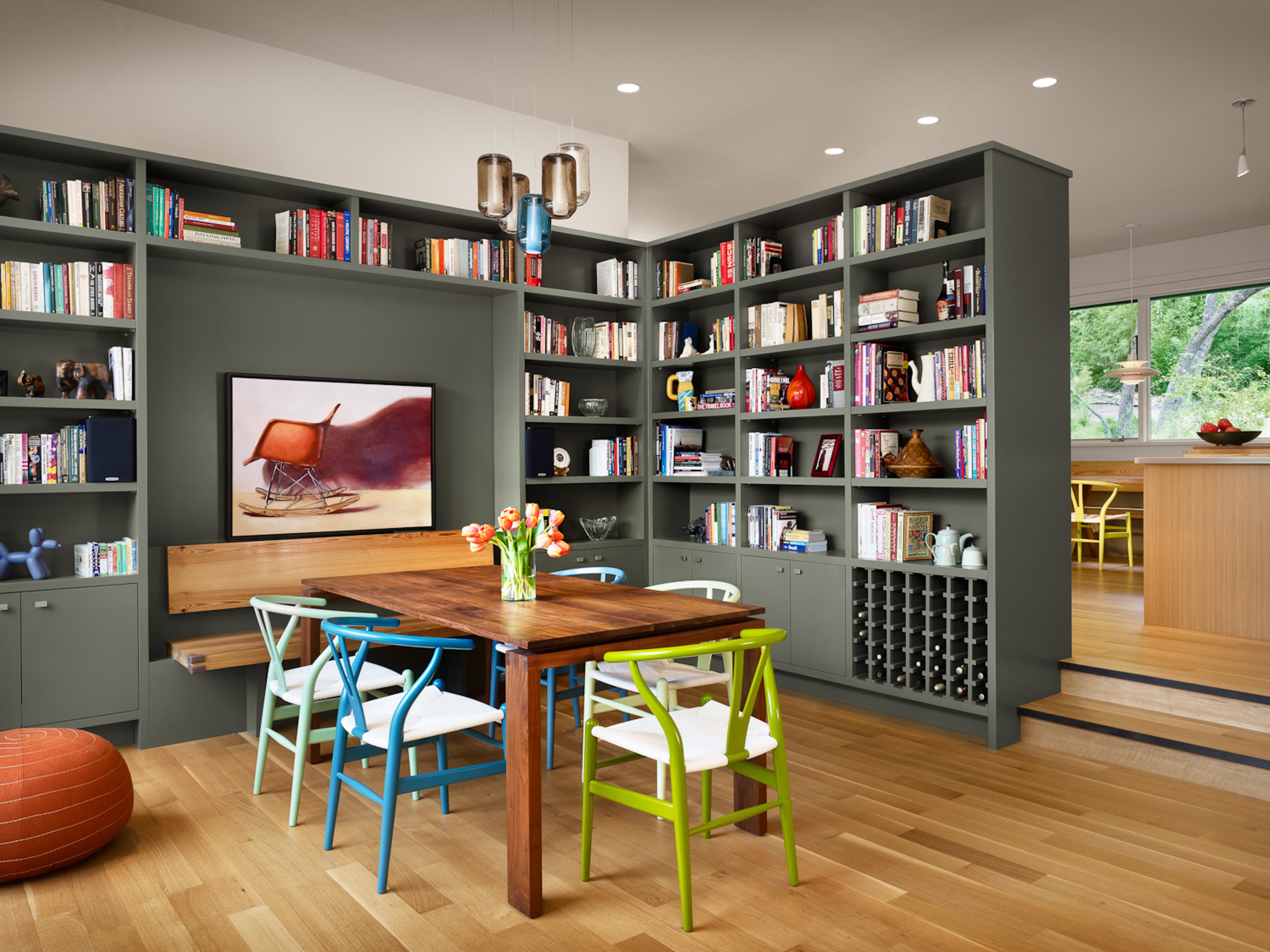 A modern dining area features a wooden table surrounded by colorful chairs, next to a bookshelf filled with books and decorative items.