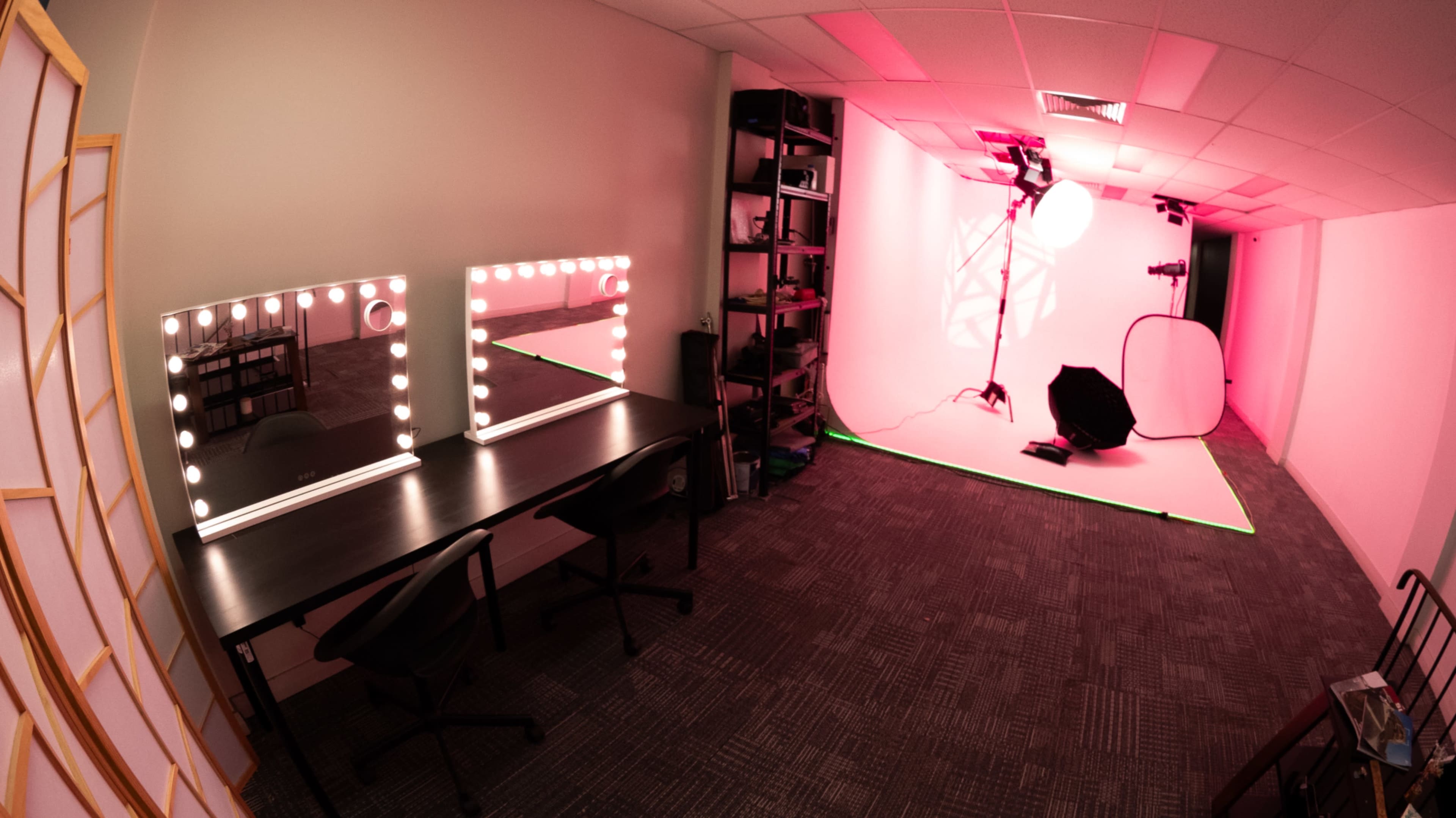 The image shows a brightly lit studio space with two large mirrors surrounded by light bulbs, a black table with chairs, and a photo setup in the background.