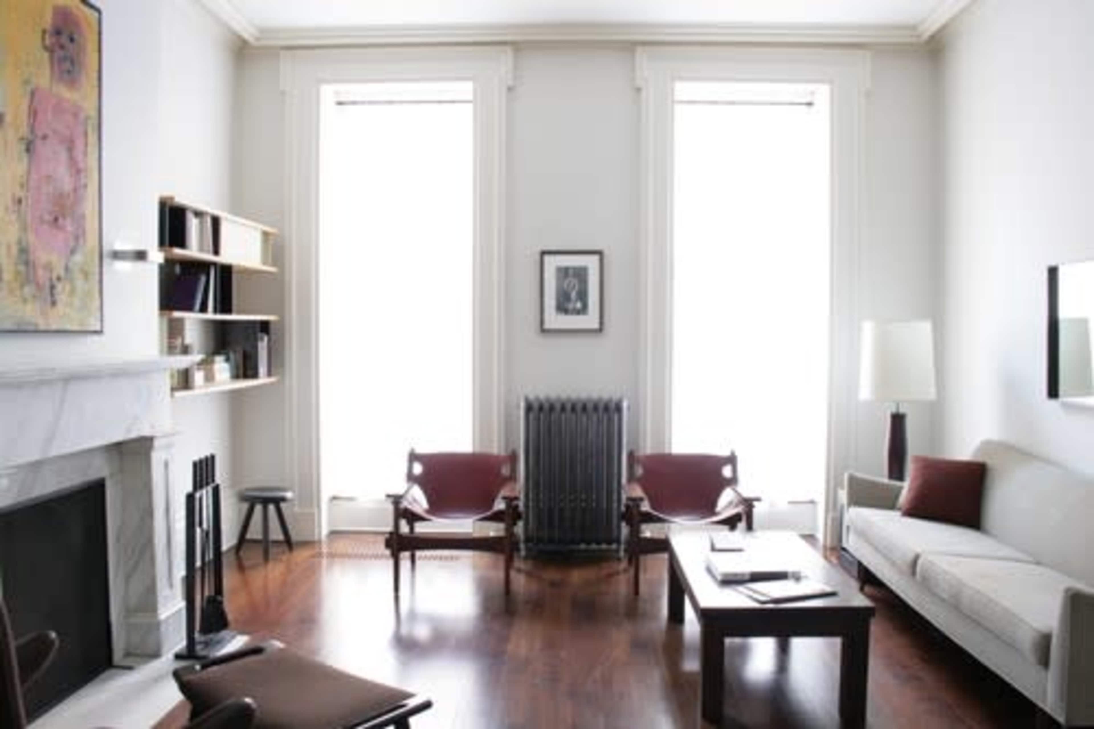 The room features a minimalist design with a light-colored sofa, two red chairs, a wooden coffee table, and large windows that allow natural light to enter.