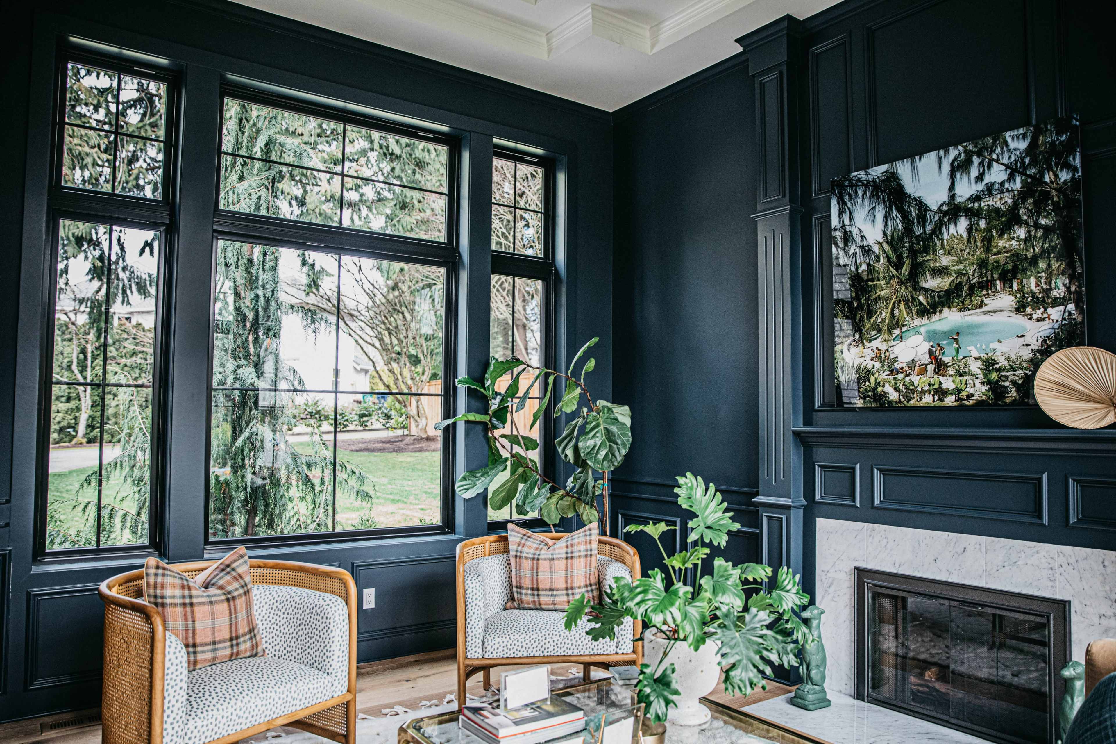 A modern living room features two wicker chairs with plaid cushions, a leafy plant, and large windows that overlook a lush outdoor view.