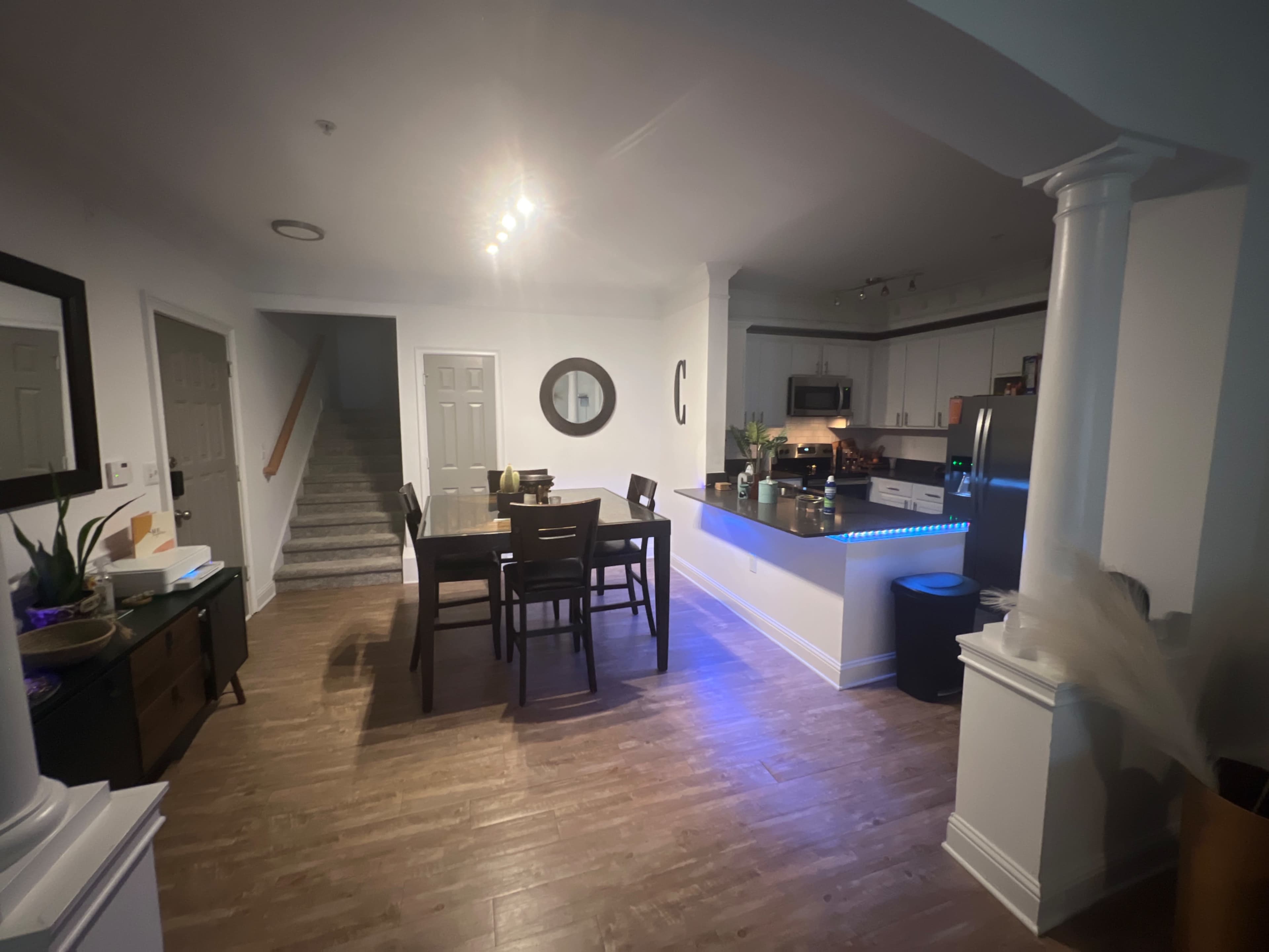 The image shows a dining area connected to a kitchen, featuring a wooden table with chairs and a staircase in the background.