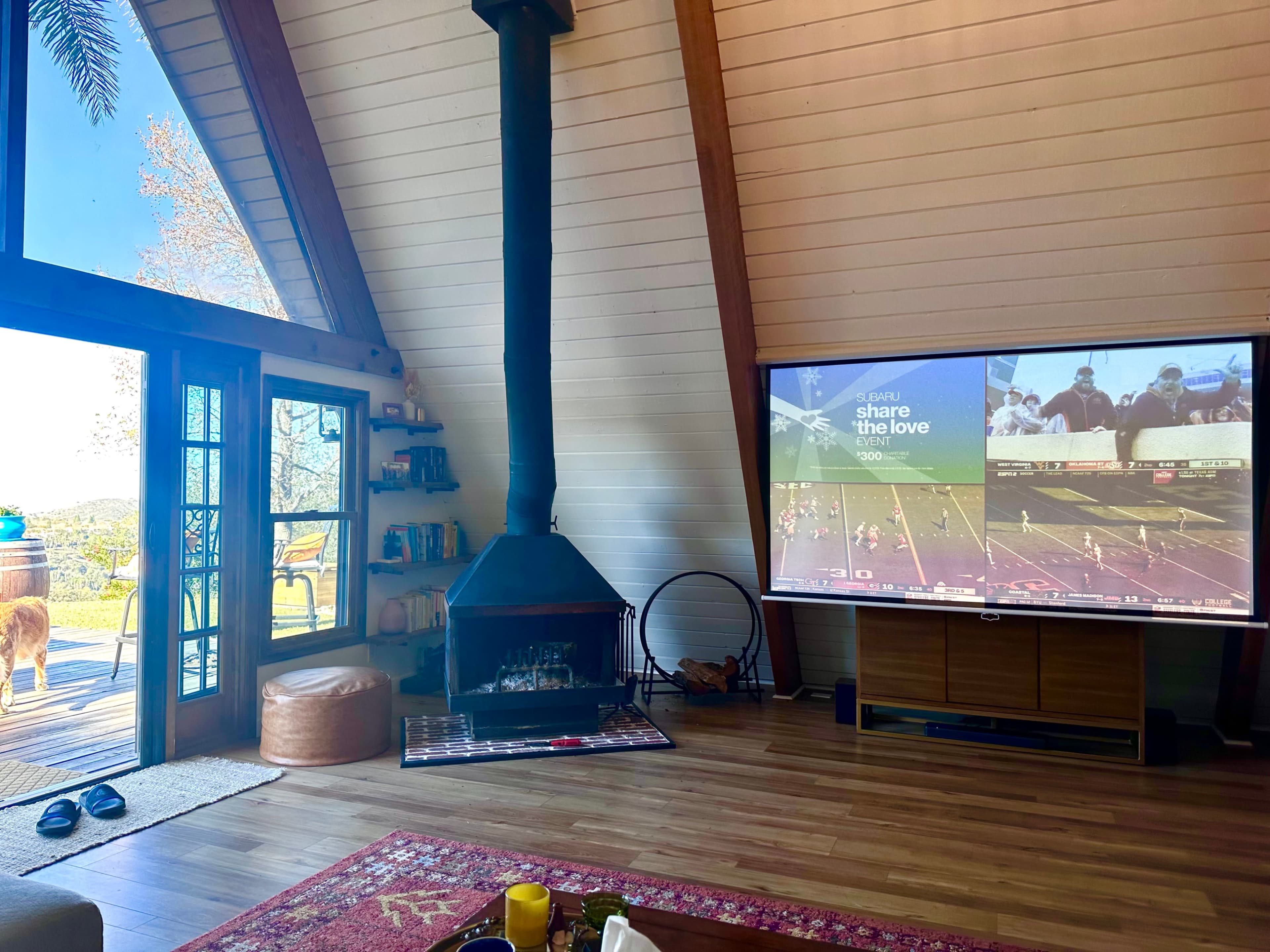 A spacious living room with a large screen displaying a sports game, a fireplace, and a view of the outdoors through large glass doors.