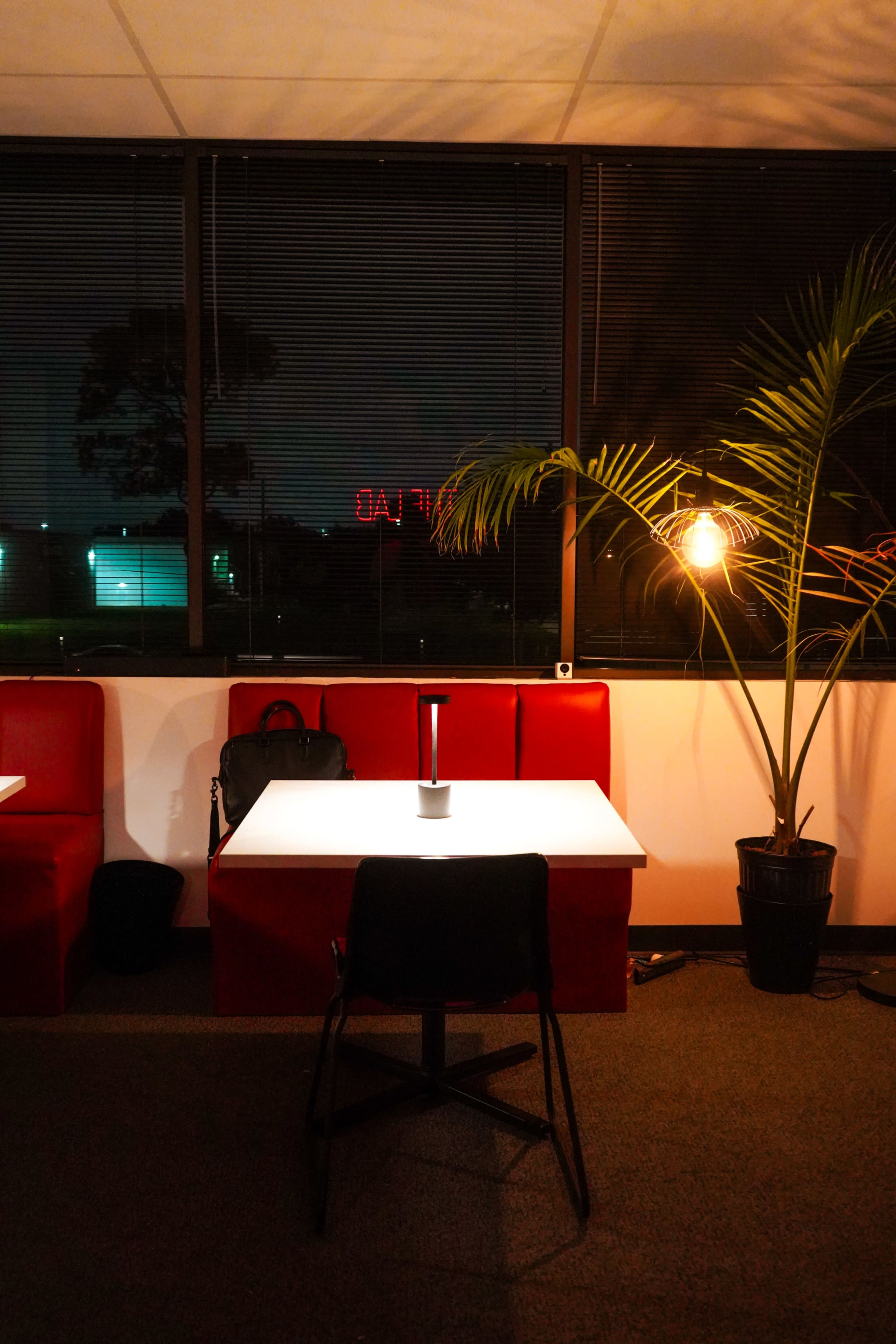 A dimly lit cafe with red booths, a potted palm tree, and a small lamp on a white table against a window with night views.