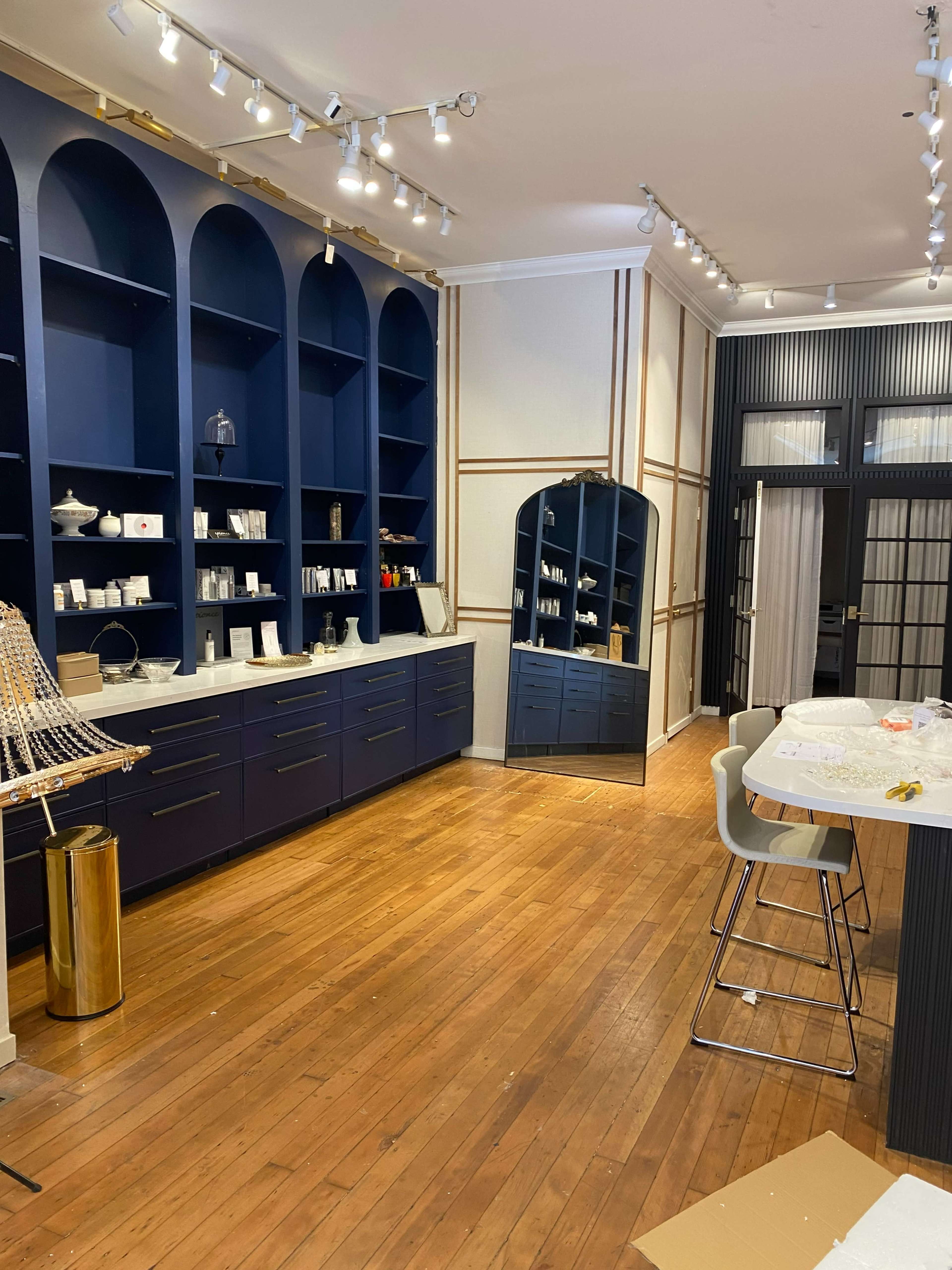 The image shows a stylish retail space featuring wooden flooring, navy blue shelving along the walls, a large mirror, and a countertop with bar stools.