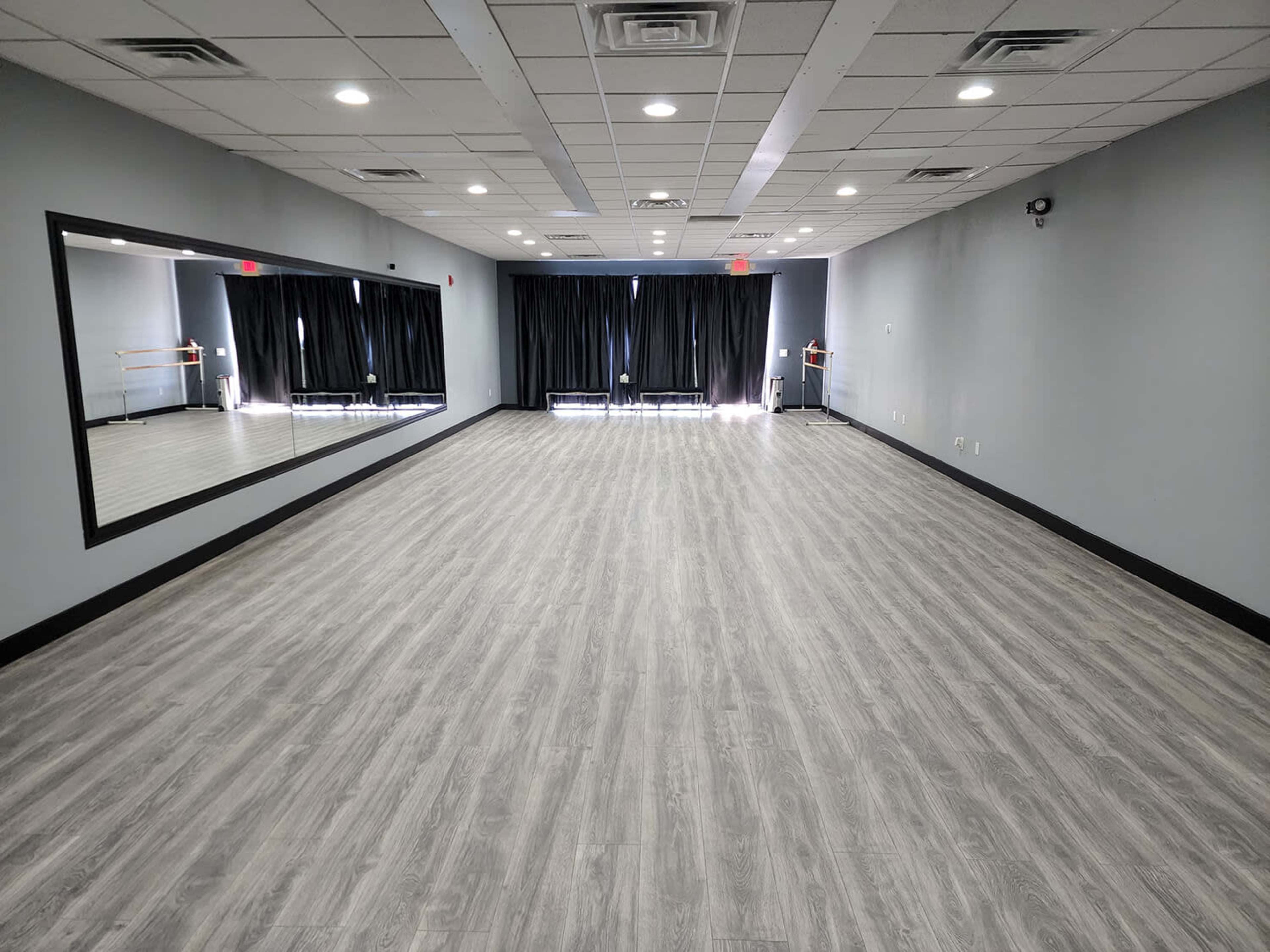 A spacious dance studio with gray walls, a large mirror on one side, and black curtains covering the windows.