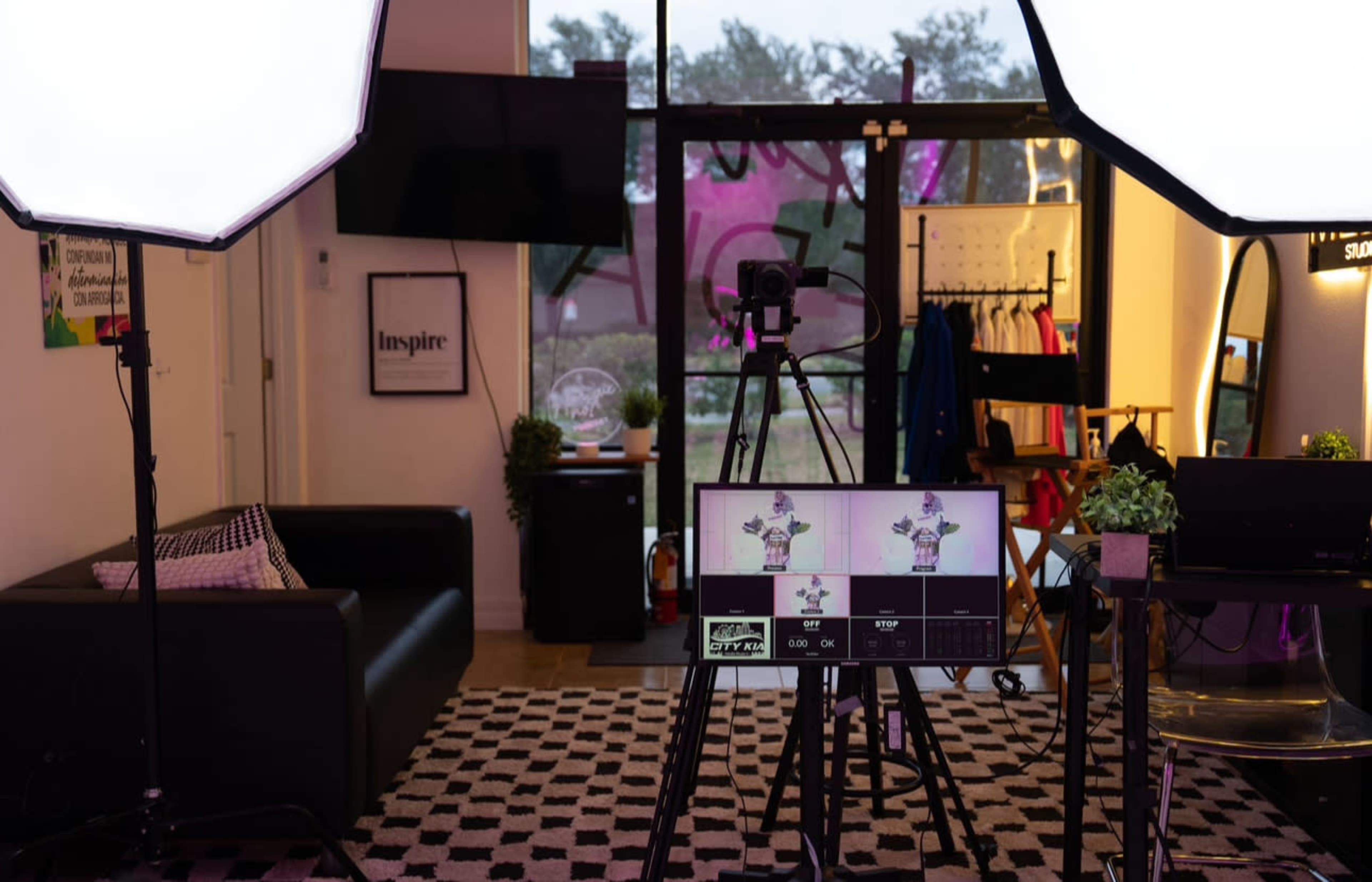 The image shows a modern studio setup with lighting equipment, a camera on a tripod, and a large monitor displaying a digital interface, set within a space featuring a black couch and colorful clothing in the background.