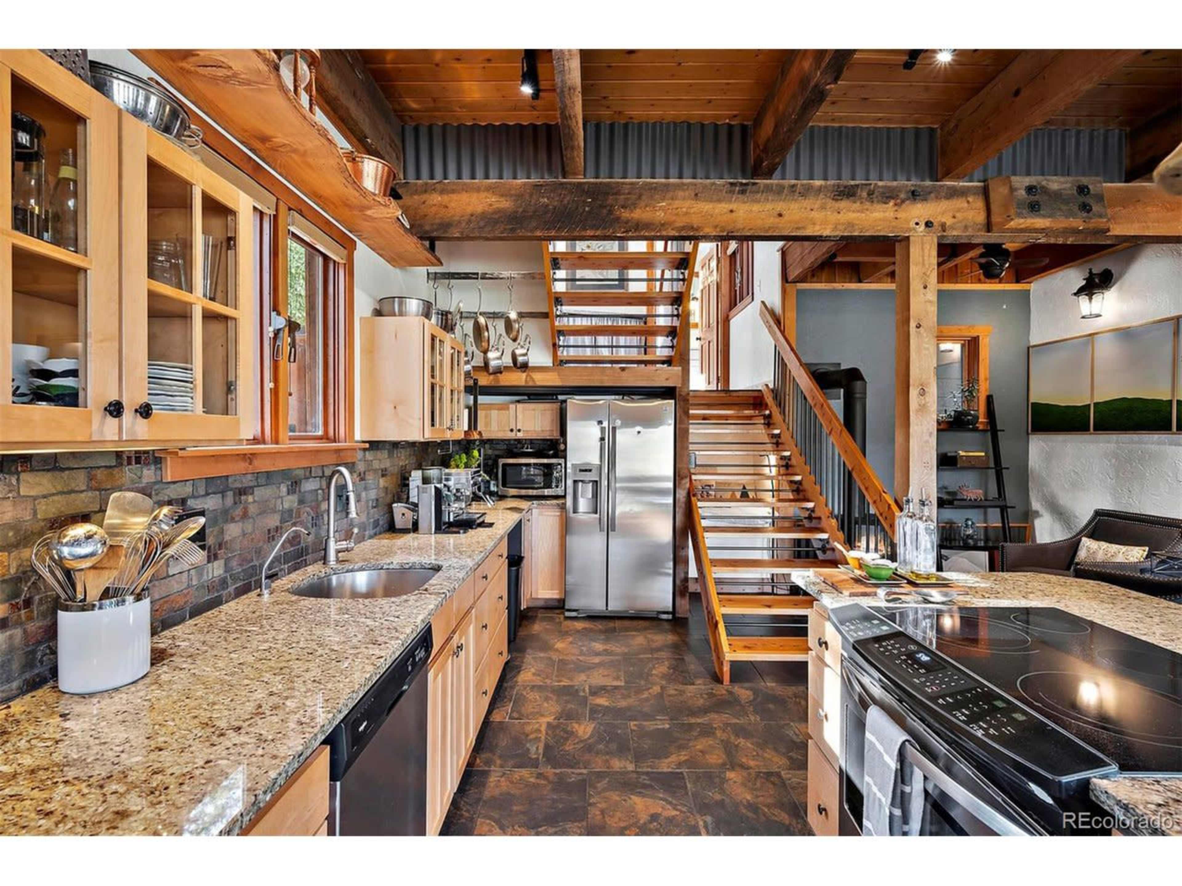A modern kitchen with wooden cabinets, granite countertops, and stainless steel appliances, leading to a staircase in a rustic setting.