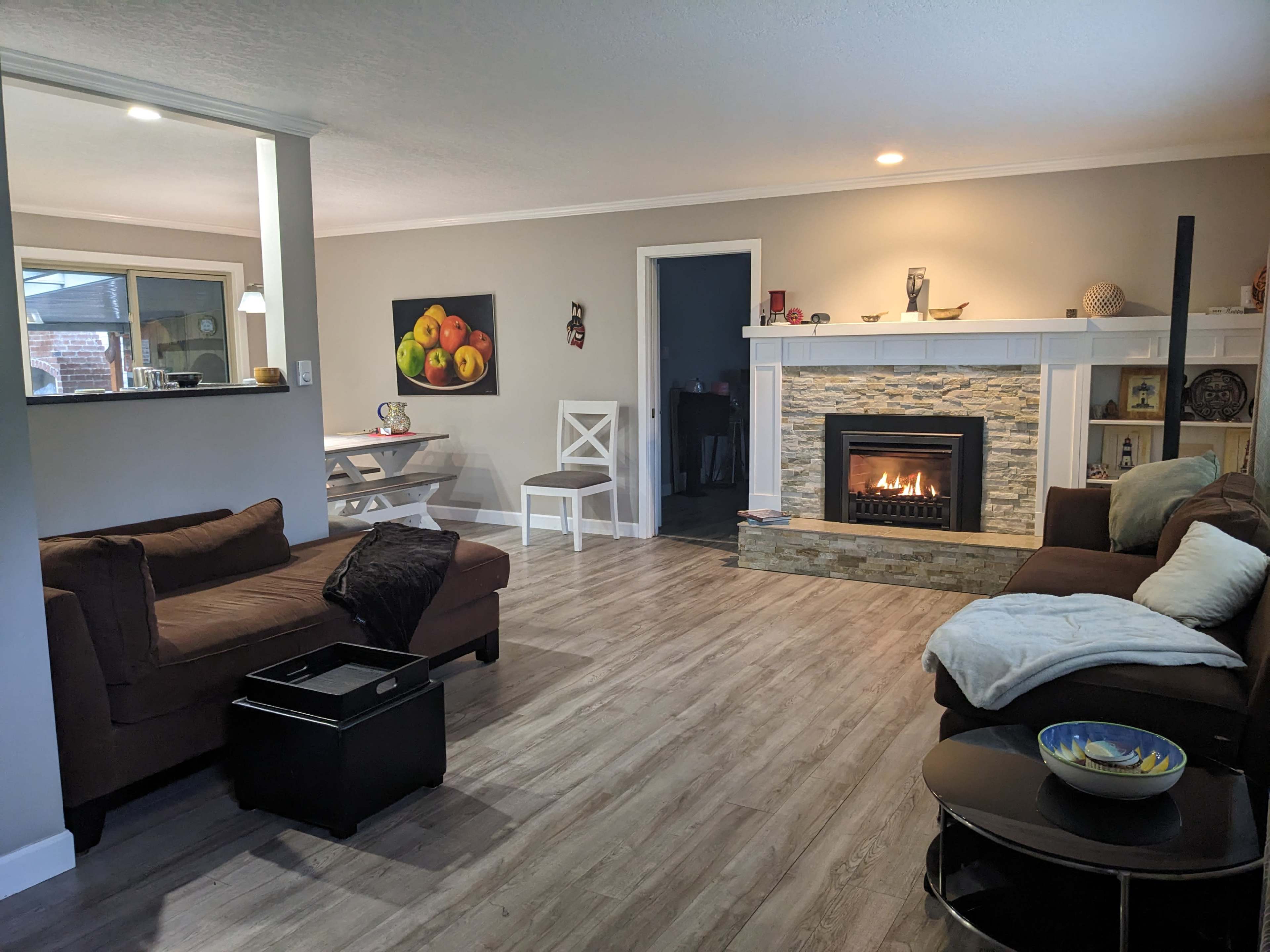 The image shows a living room with a stone fireplace, a brown sectional sofa, a white dining table, and a decorative bowl on a coffee table.