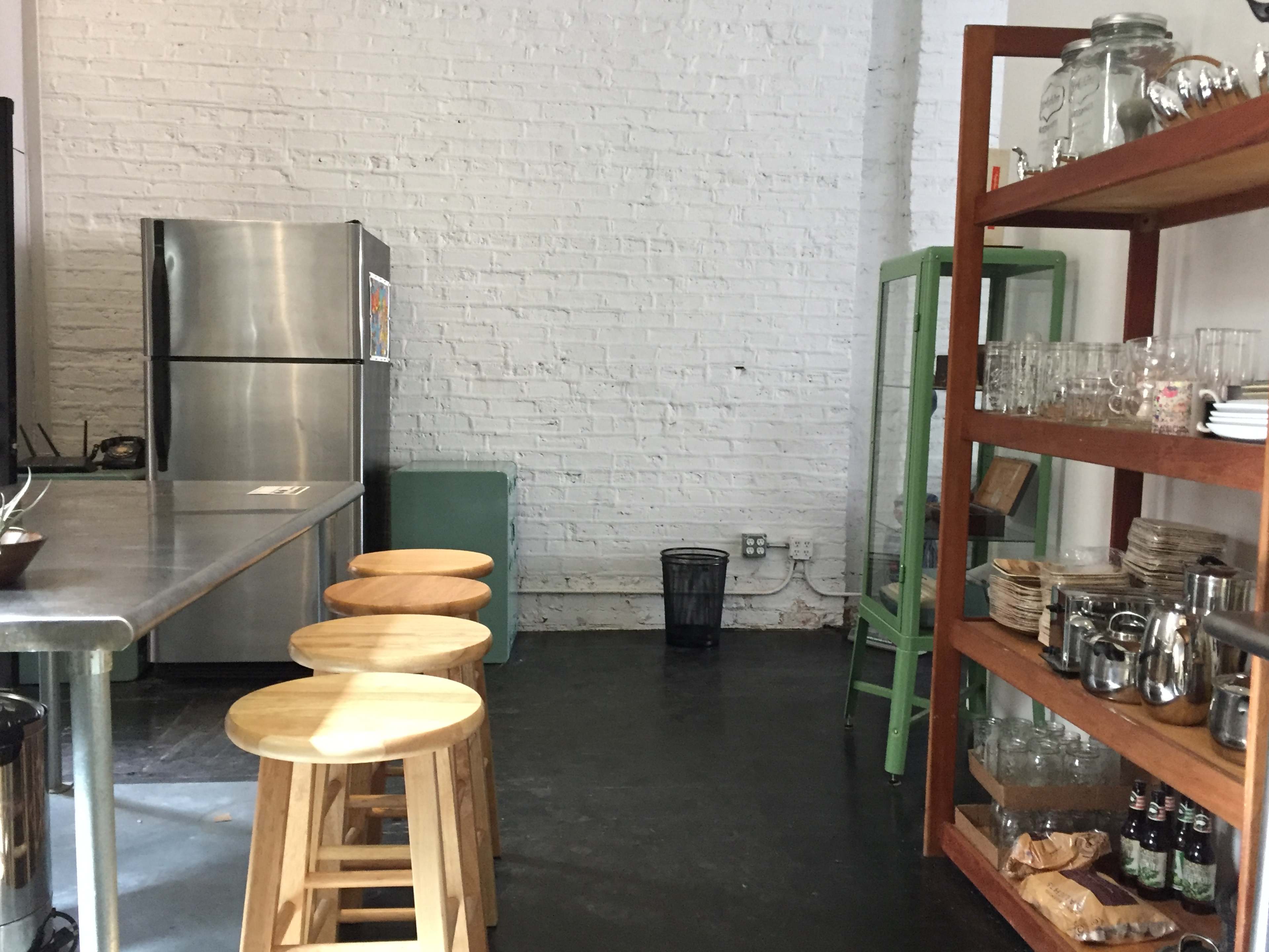 A kitchen area with a stainless steel countertop, wooden stools, a refrigerator, shelves filled with glassware, and a green cabinet against a white brick wall.