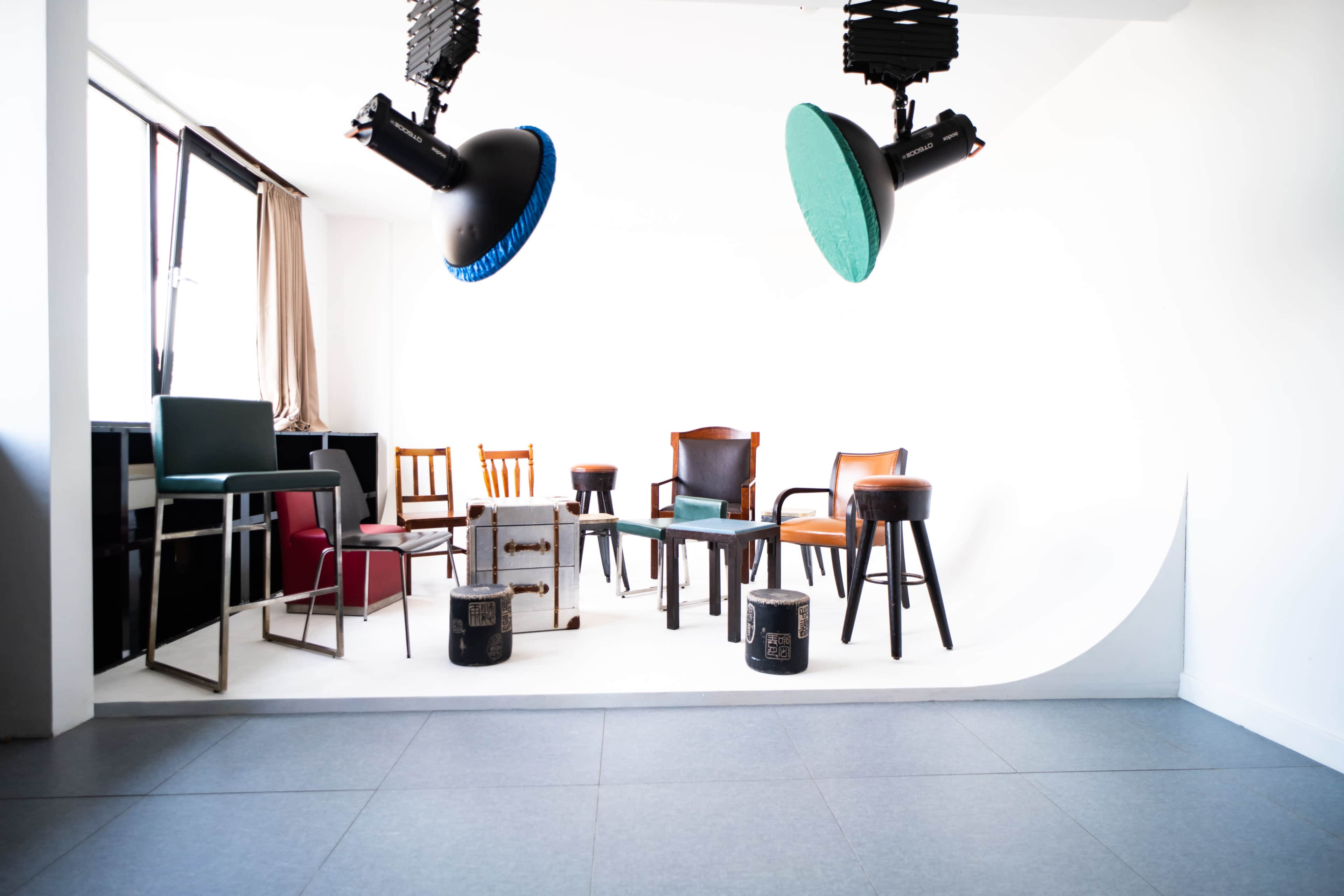 The image shows a minimalist photo studio setup featuring various chairs and stools arranged against a curved white backdrop, with studio lights hanging above.