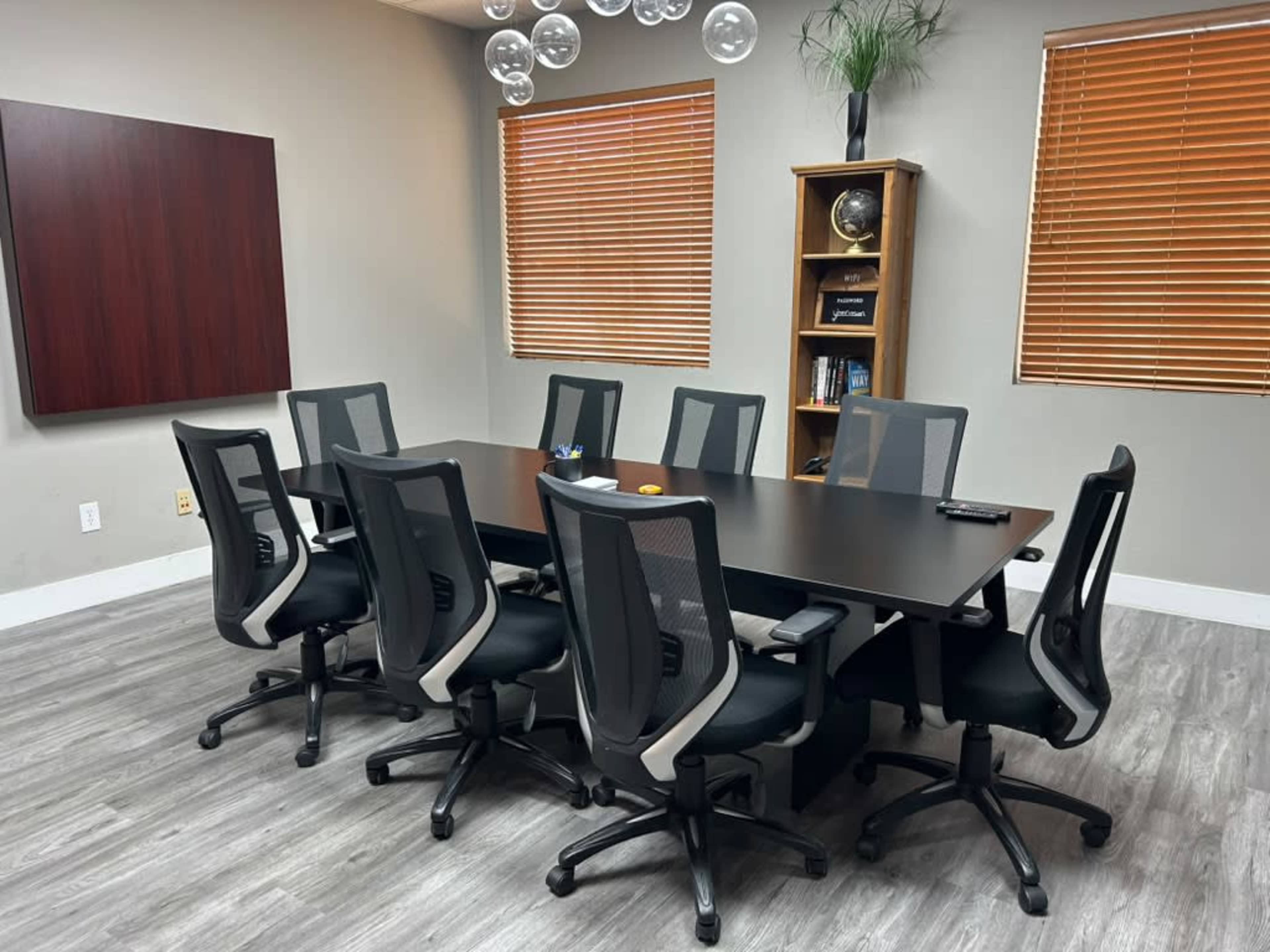 The image shows a conference room with a long table surrounded by eight ergonomic chairs, large windows with wooden blinds, and a bookshelf in the corner.