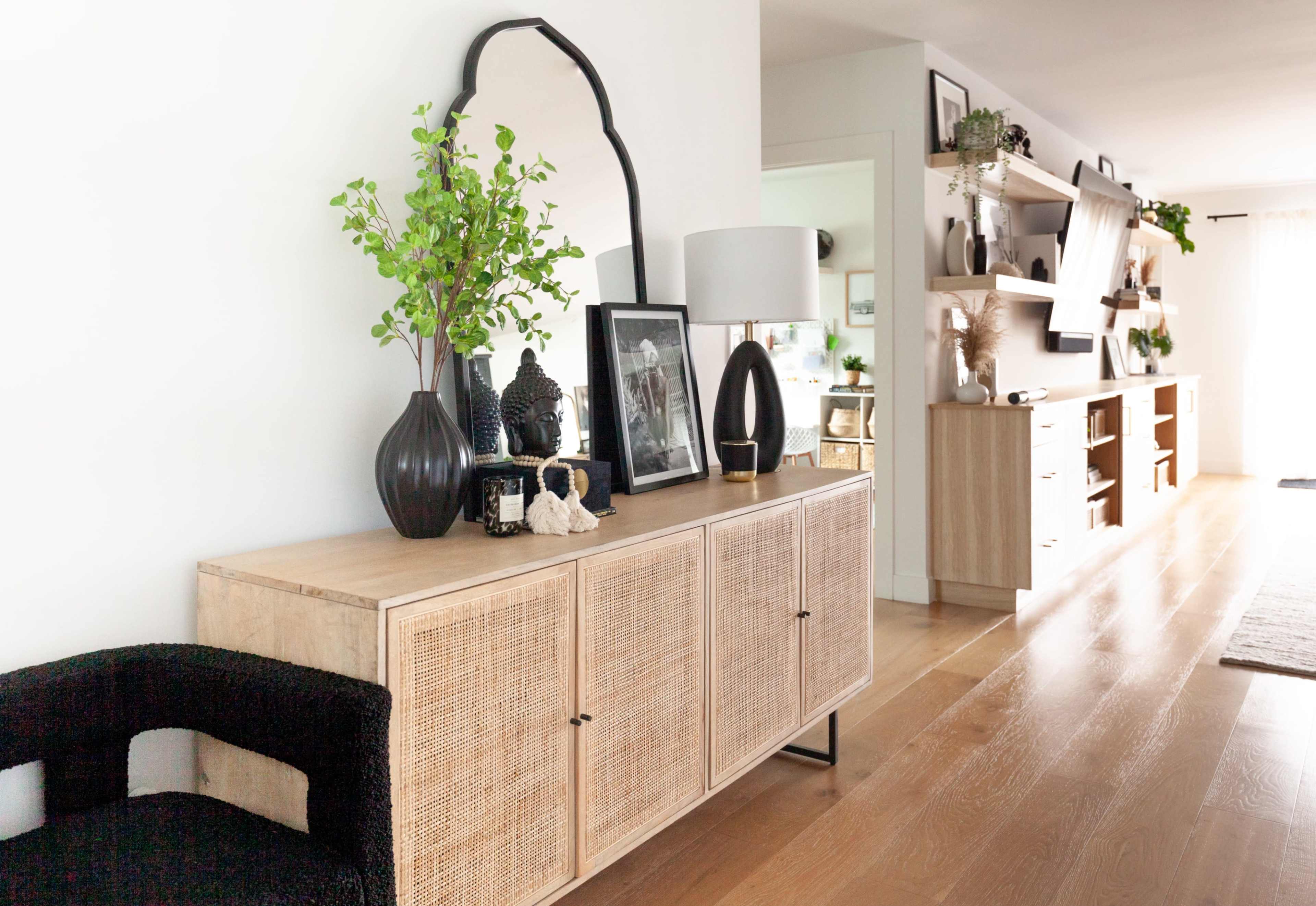A wooden sideboard with decorative items and plants stands against a white wall in a bright, minimalist room that features wooden flooring and shelves.