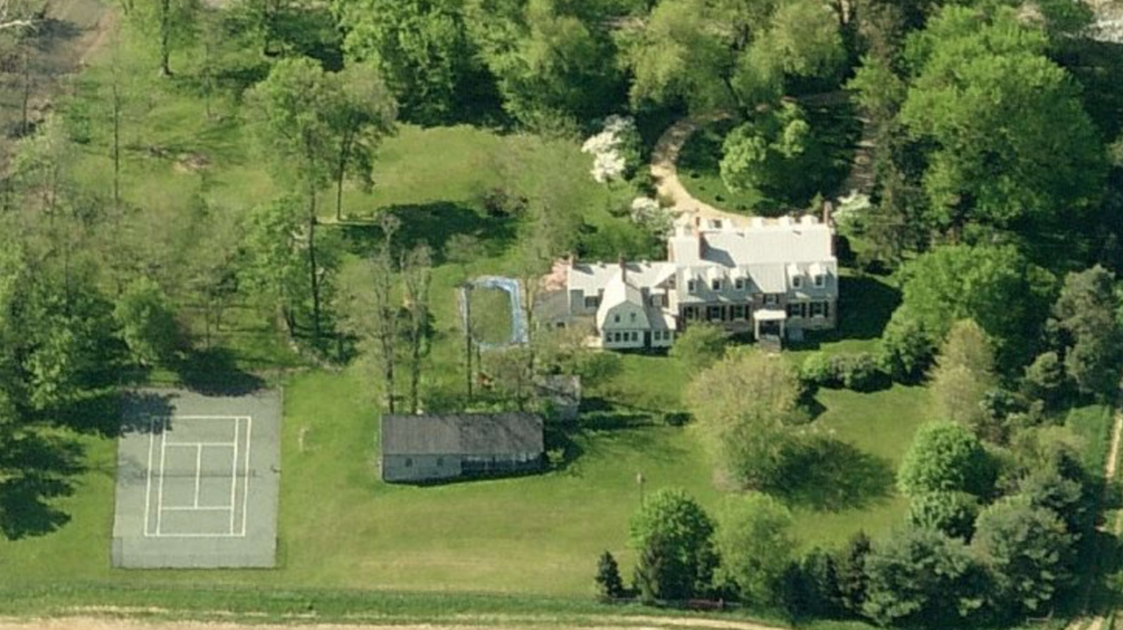 An aerial view shows a large house surrounded by trees, a swimming pool, and a tennis court nearby.