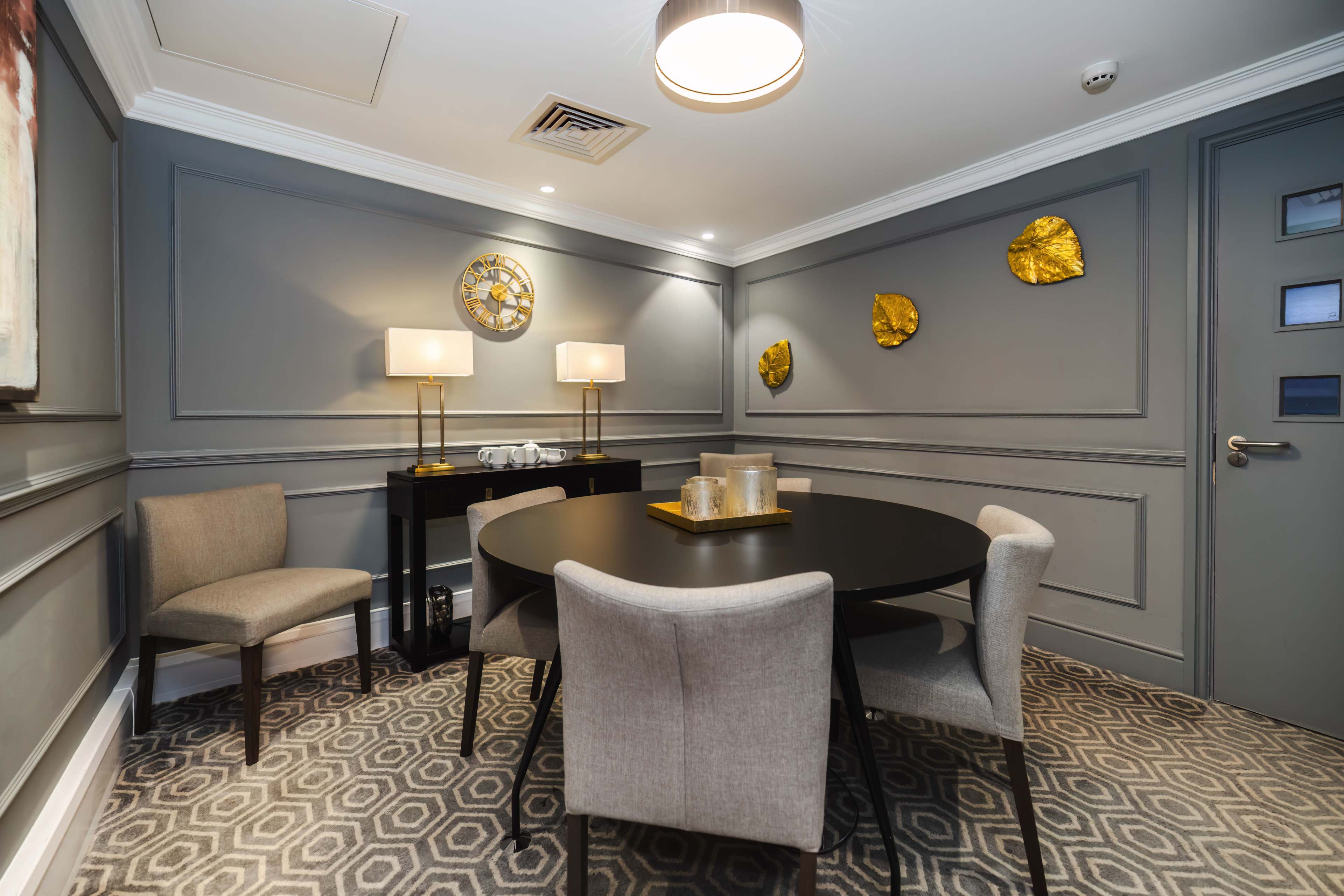 A small dining area featuring a round black table surrounded by four light-colored chairs, a console table with decorative items, and gold wall decor against gray walls.