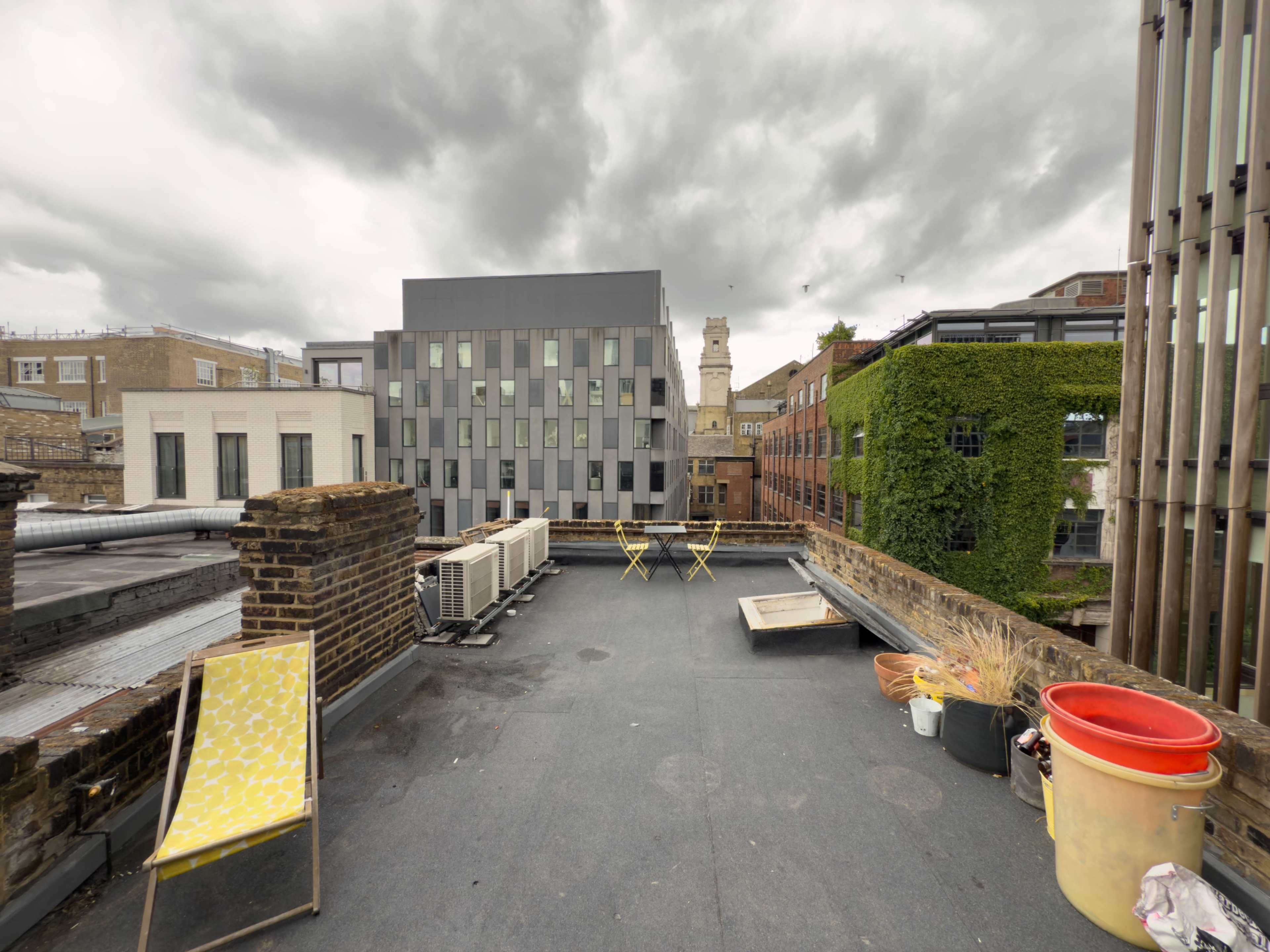 The image shows a rooftop with two yellow chairs, a red container, and buildings in the background under a cloudy sky.