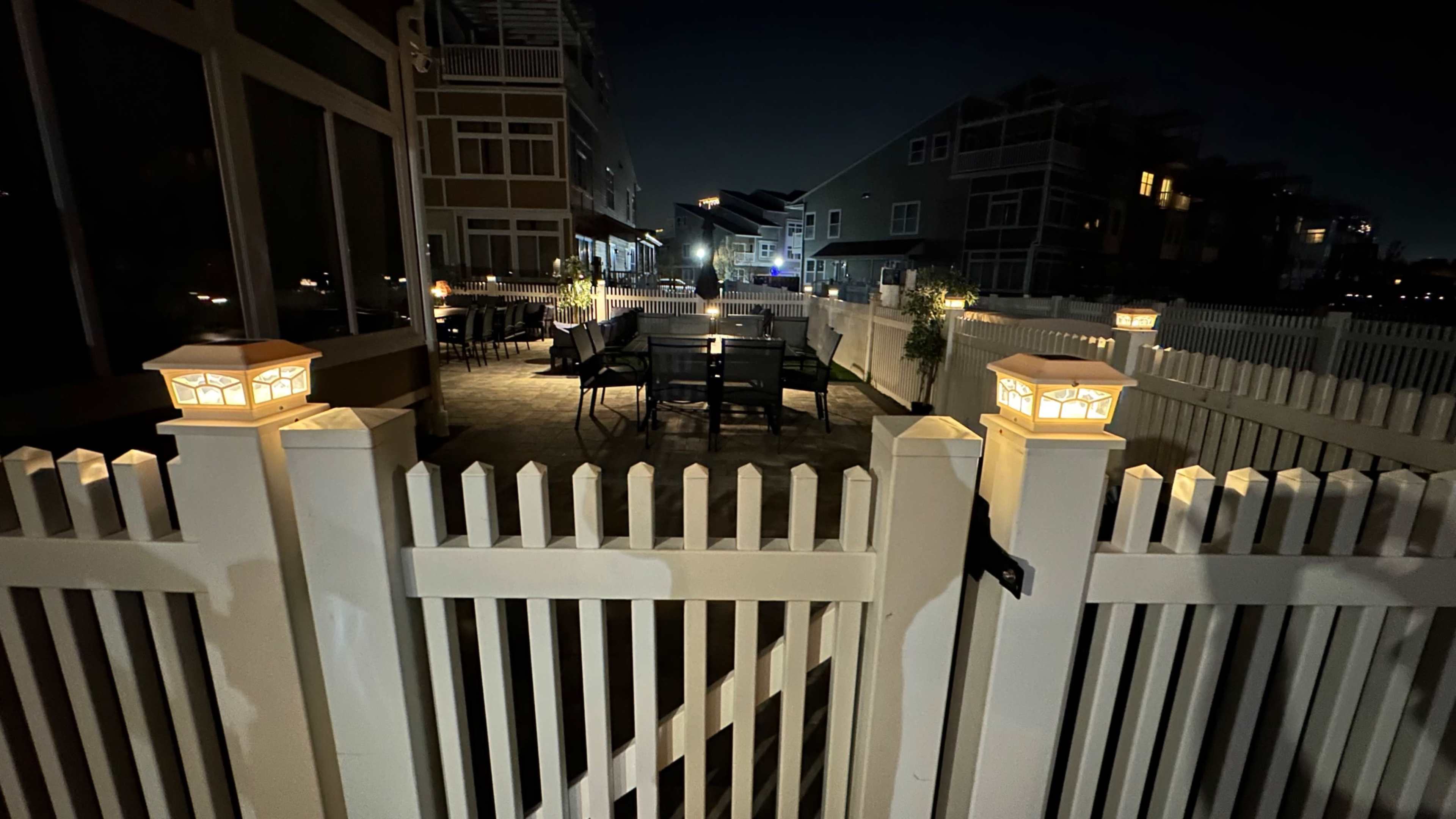 The image shows a well-lit patio area enclosed by a white picket fence, with tables and chairs arranged under soft lighting at night.