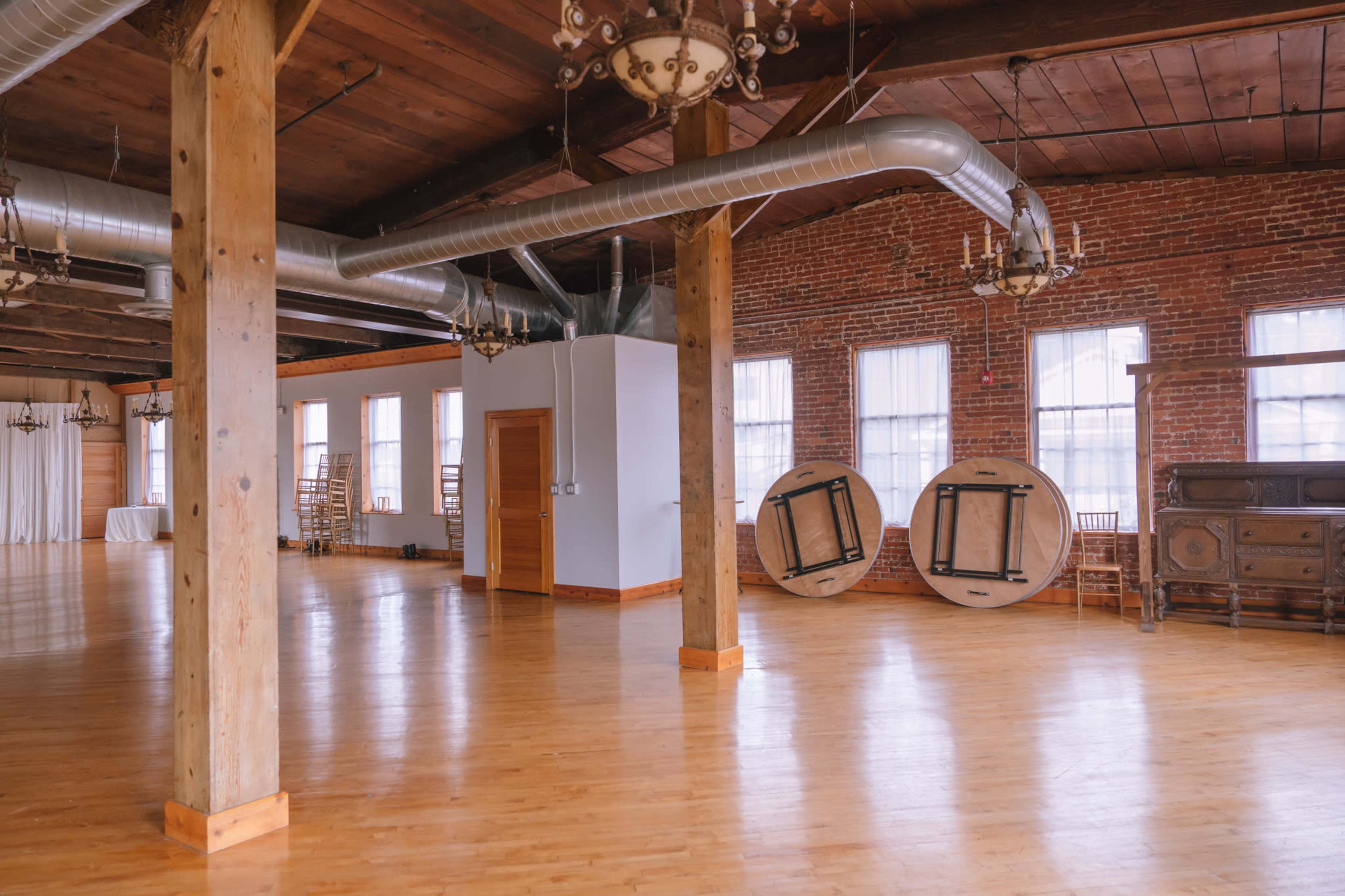 The image shows an empty event space with wooden floors, exposed brick walls, and large windows, featuring a few hanging chandeliers and two round tables leaning against a wall.