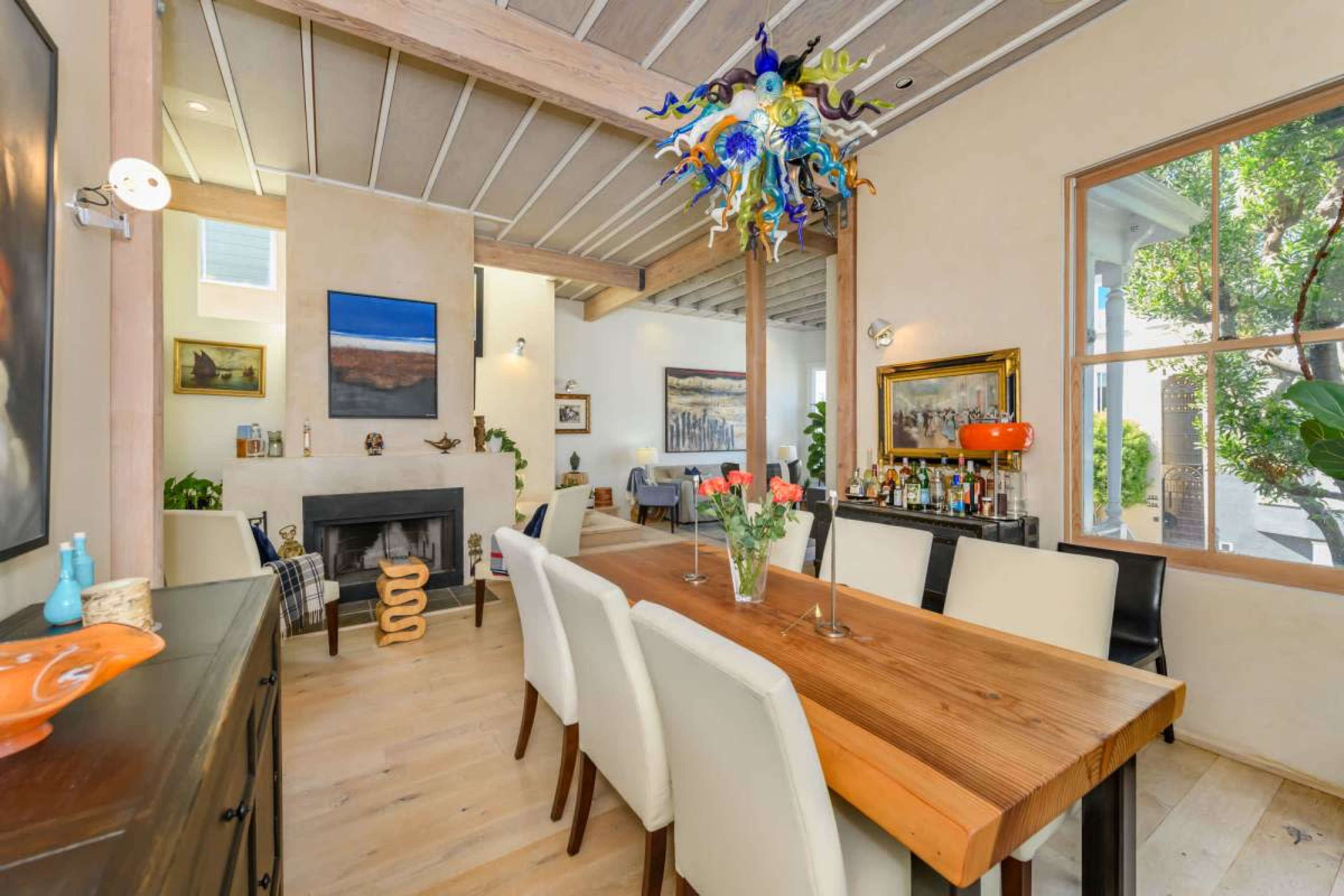 The image shows a modern dining area featuring a wooden table surrounded by white chairs, a fireplace, and colorful artwork on the walls.