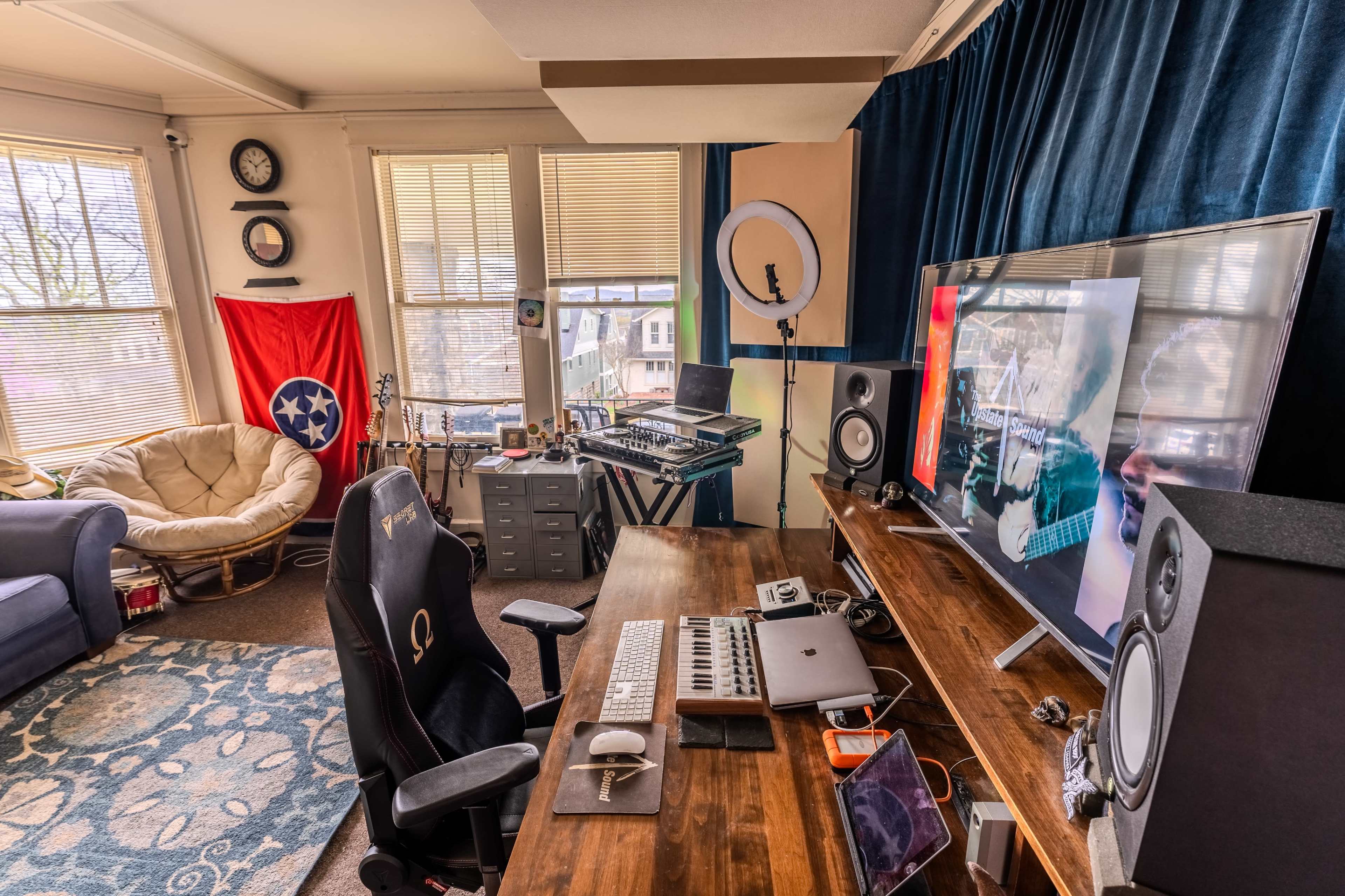 A home studio setup features a computer desk with a keyboard, speakers, and a large monitor displaying an image, surrounded by a cozy seating area and a Tennessee flag hanging on the wall.