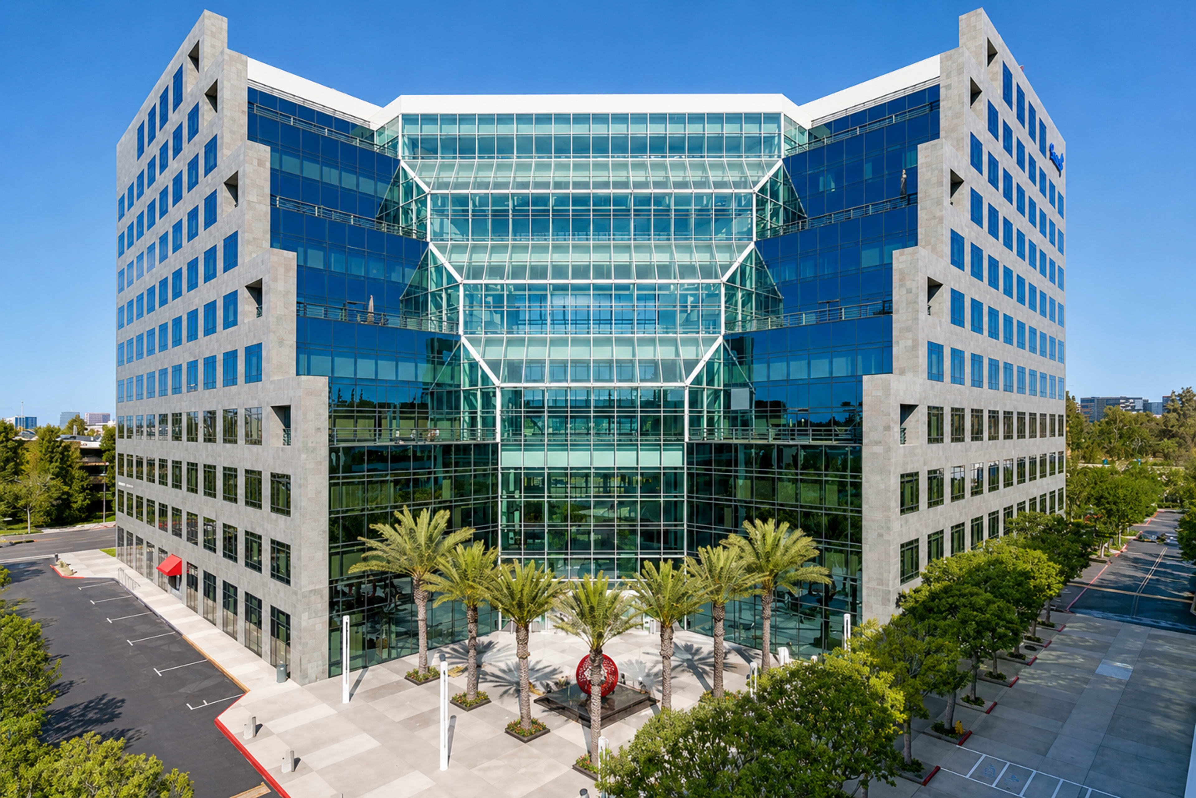 The image shows a modern, multi-story office building with large glass windows surrounded by palm trees and a clear blue sky.
