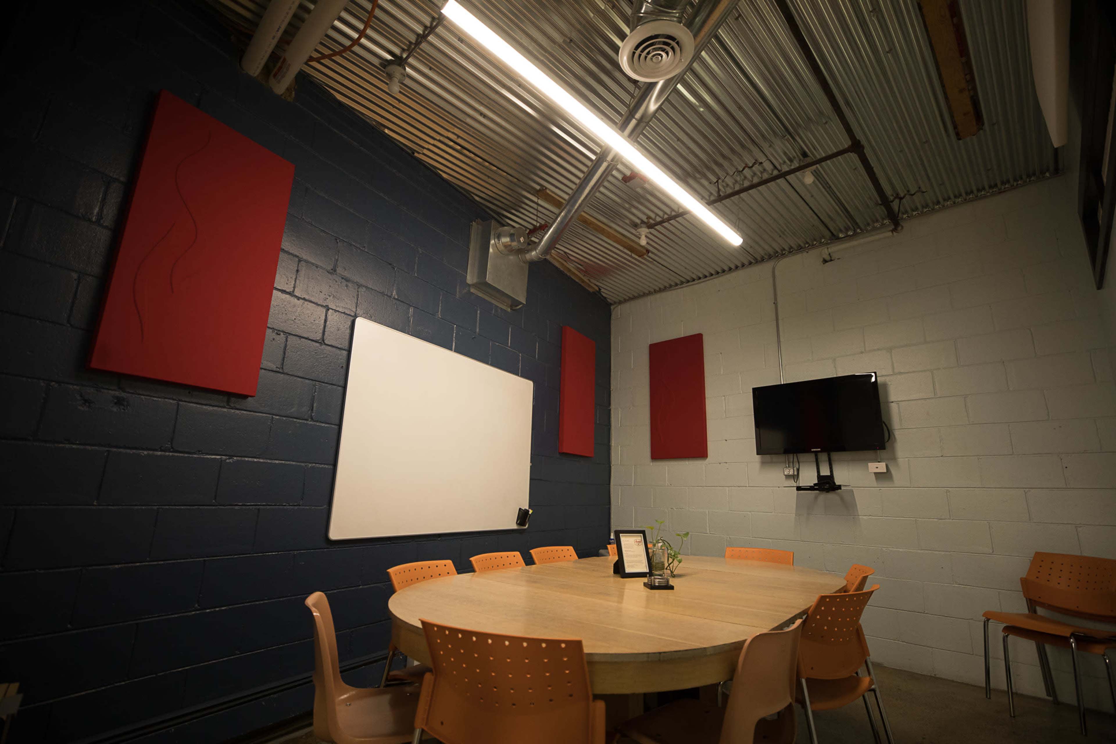 A conference room features a round table surrounded by orange chairs, with a whiteboard and wall-mounted television on opposite walls.