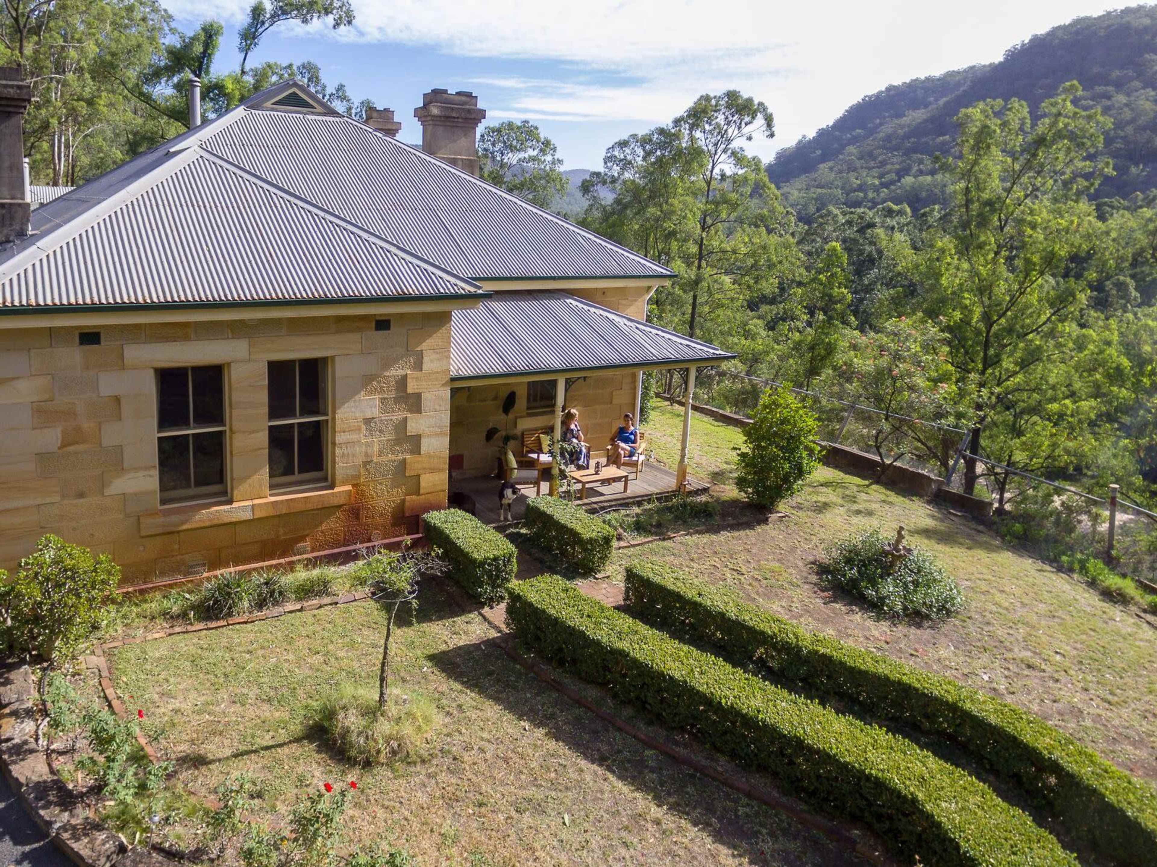 A historic sandstone house is set on a hillside surrounded by trees and manicured hedges, with people sitting on the porch.