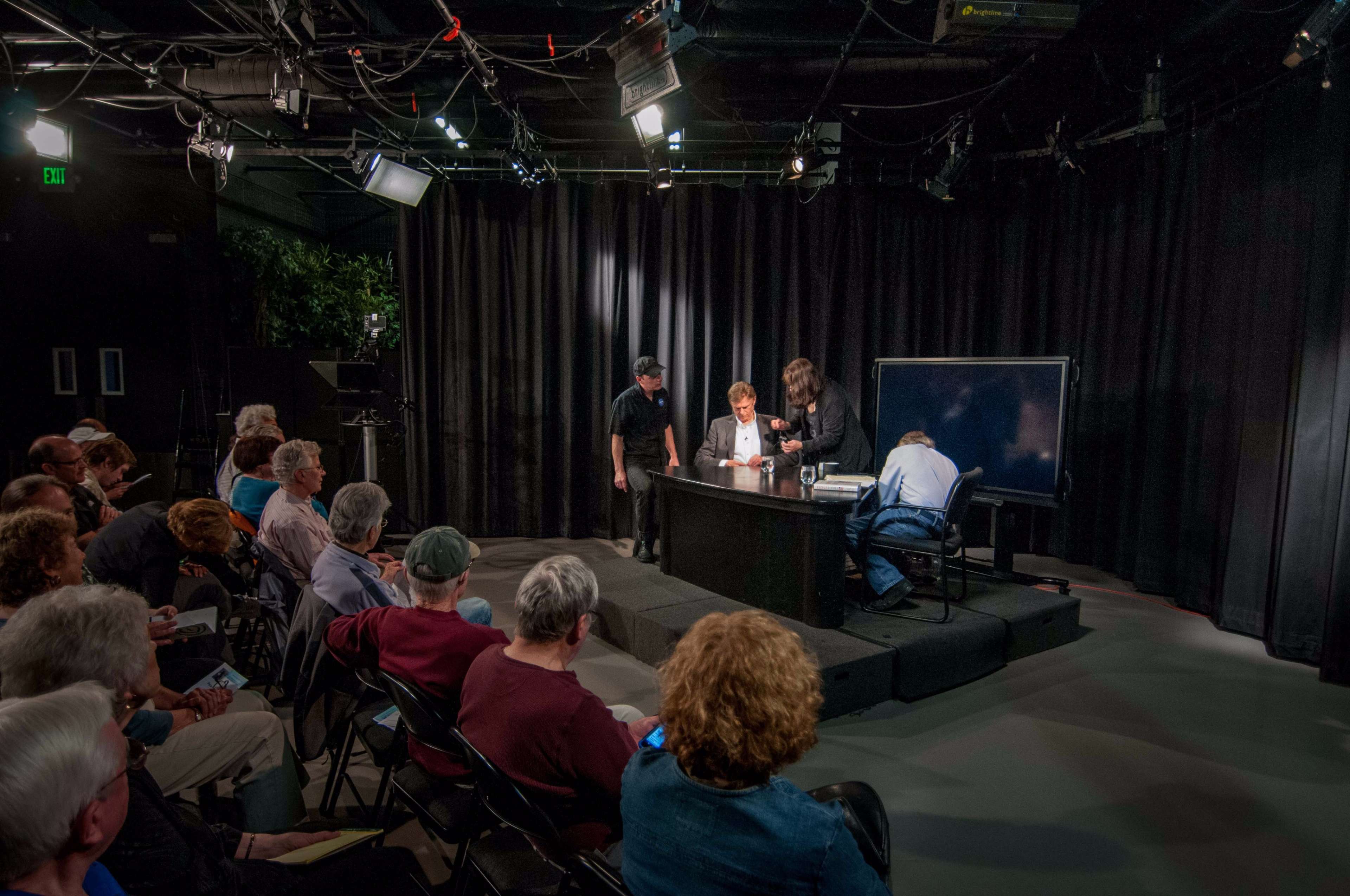 A small audience watches as four individuals prepare a presentation on a stage equipped with a black backdrop and lighting.