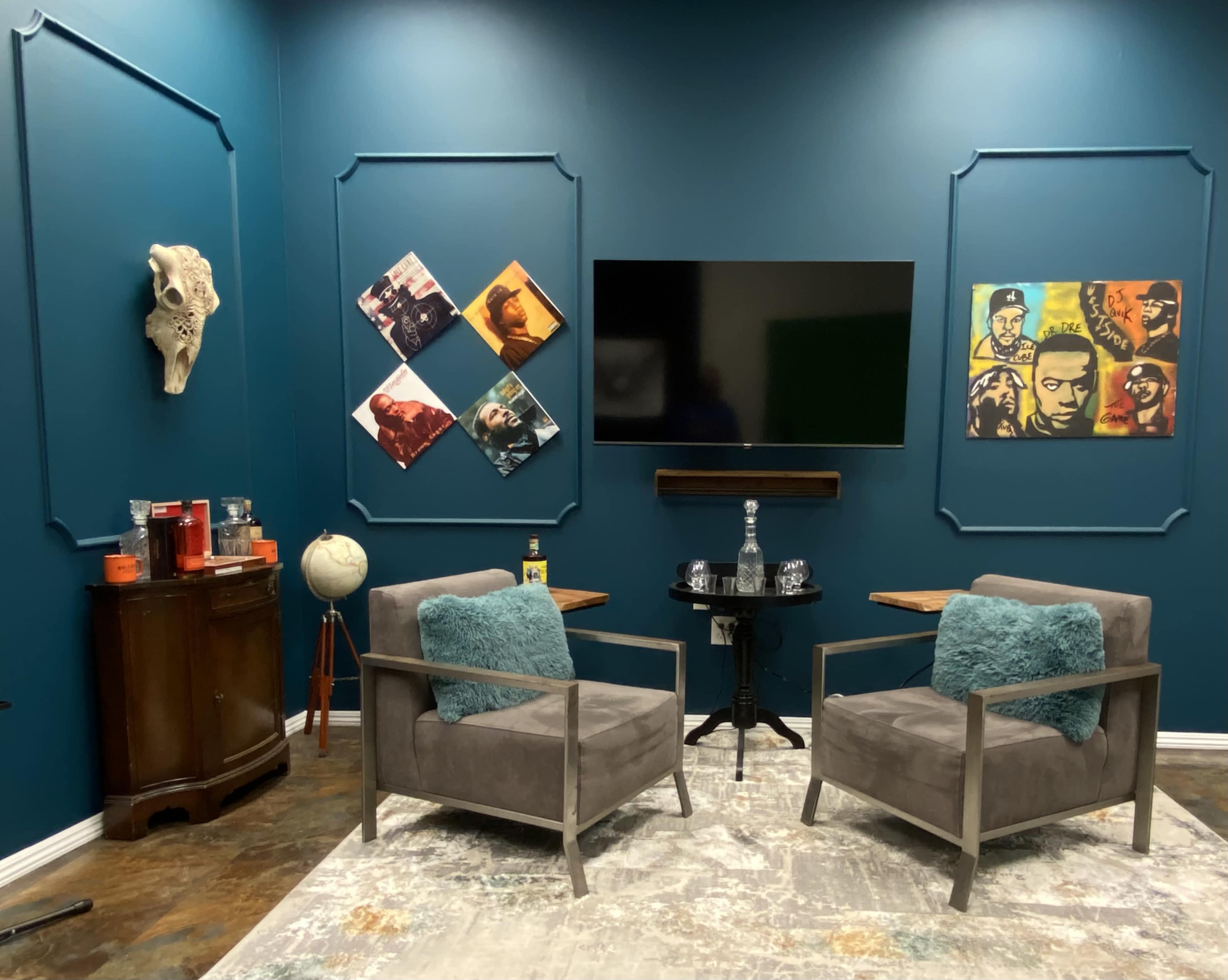 A small room with two gray chairs, a TV mounted on a teal wall, and artwork displayed alongside a globe and a wooden cabinet.