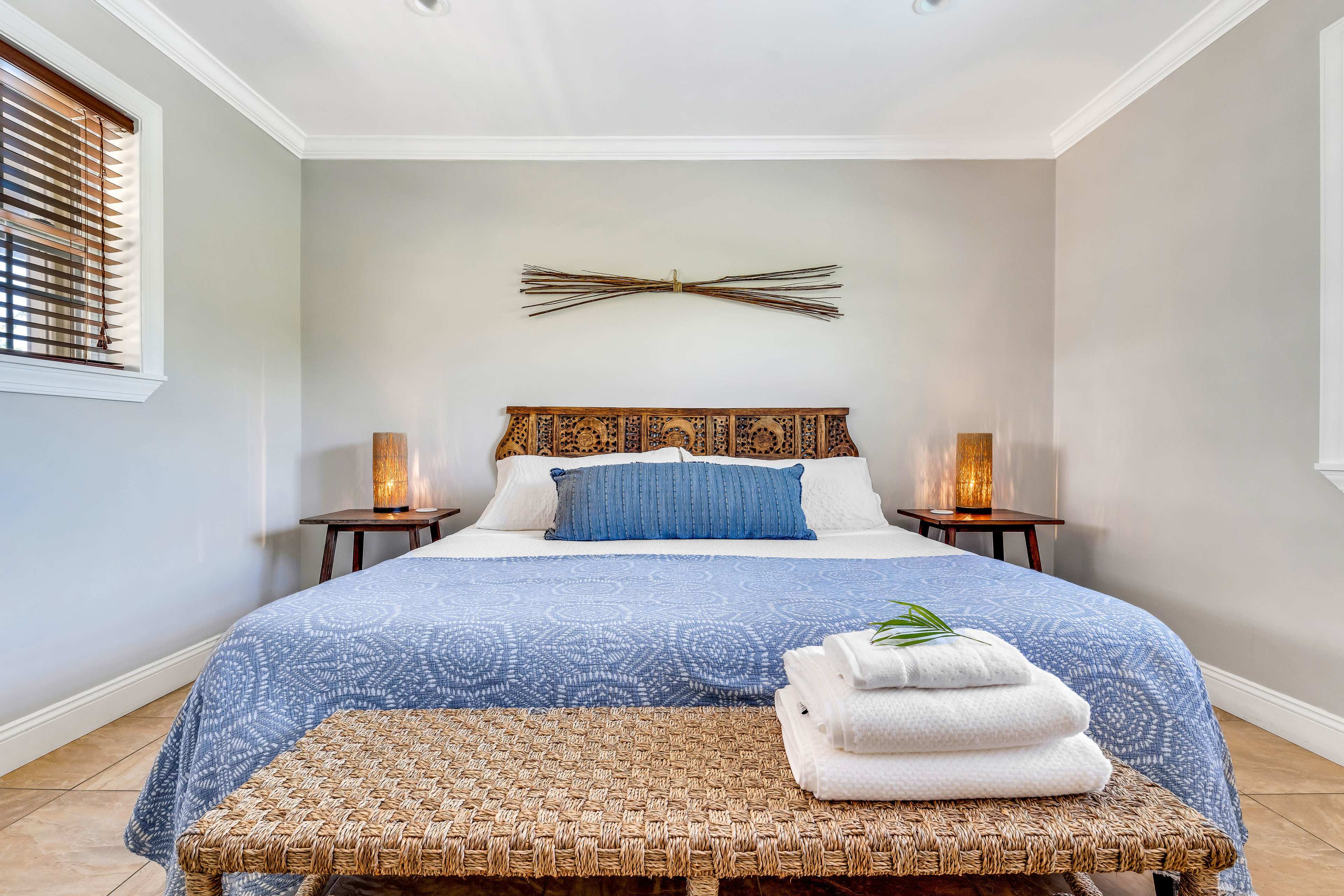 The image shows a neatly arranged bedroom with a large bed, two side tables with lamps, and a bench at the foot of the bed.