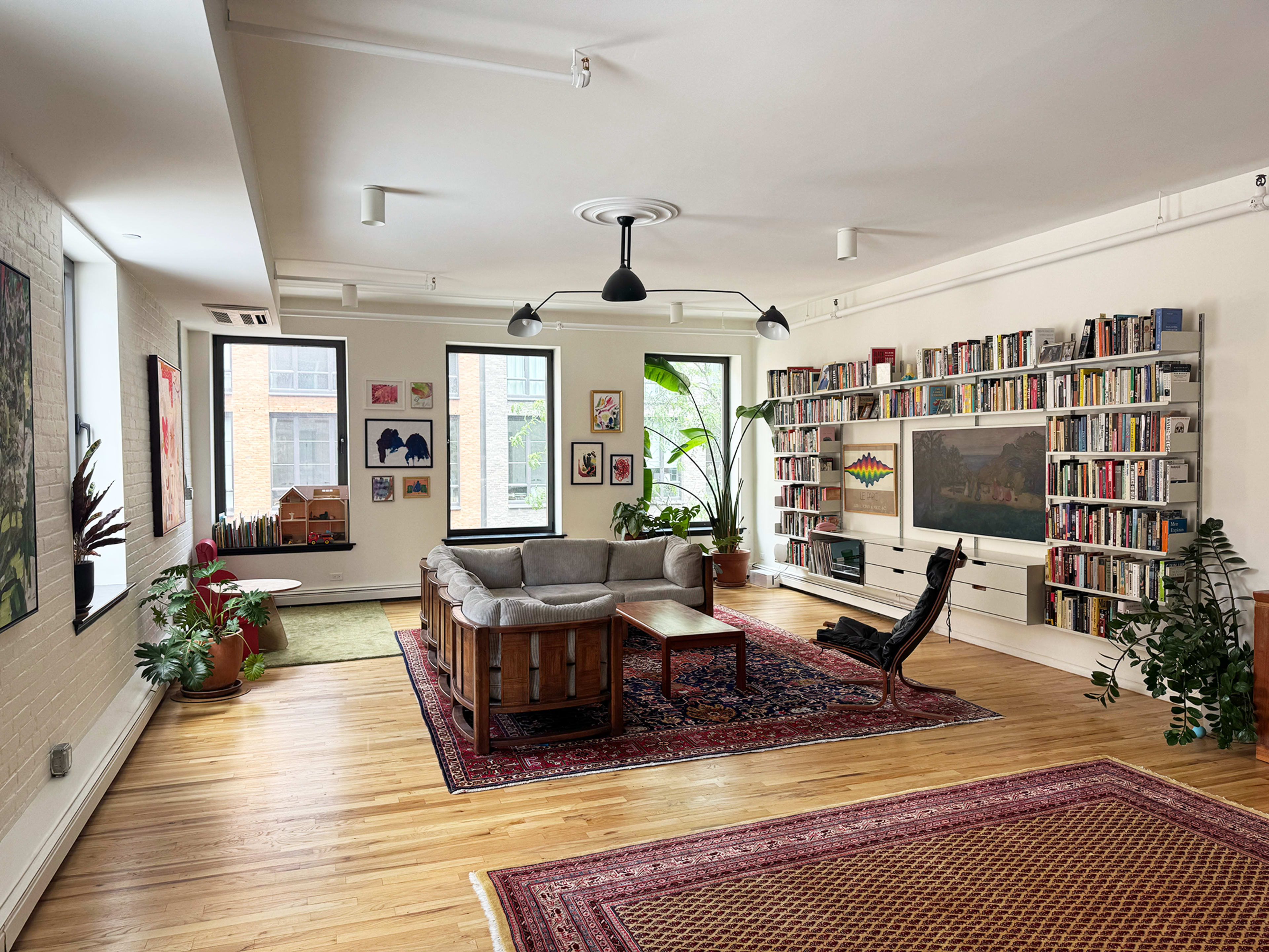 The image shows a spacious living room with a gray sectional sofa, wooden coffee table, and a bookshelf filled with books, surrounded by green plants and artwork on the walls.
