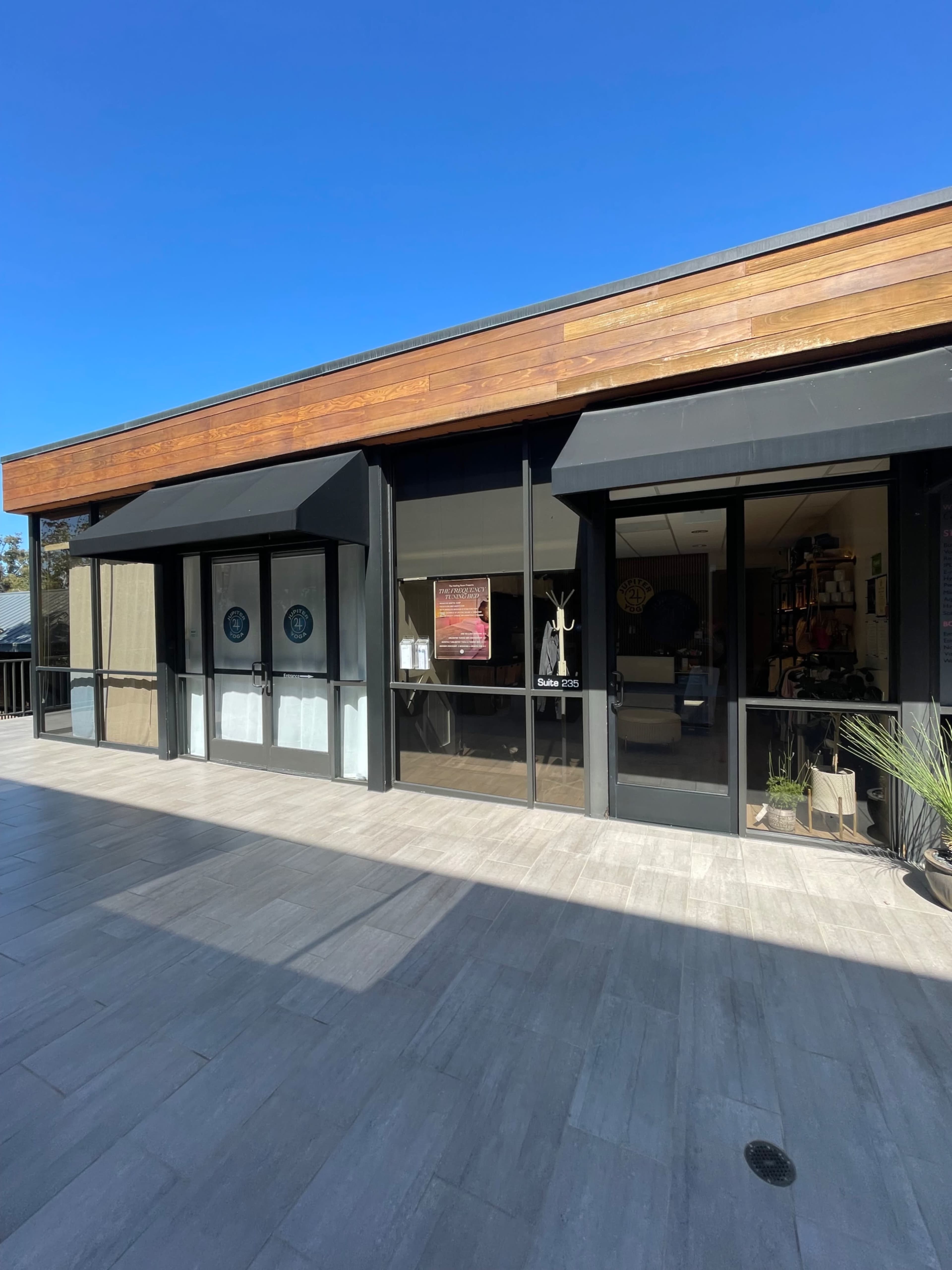The image shows a modern storefront with large glass doors and a wooden overhang, set against a clear blue sky.